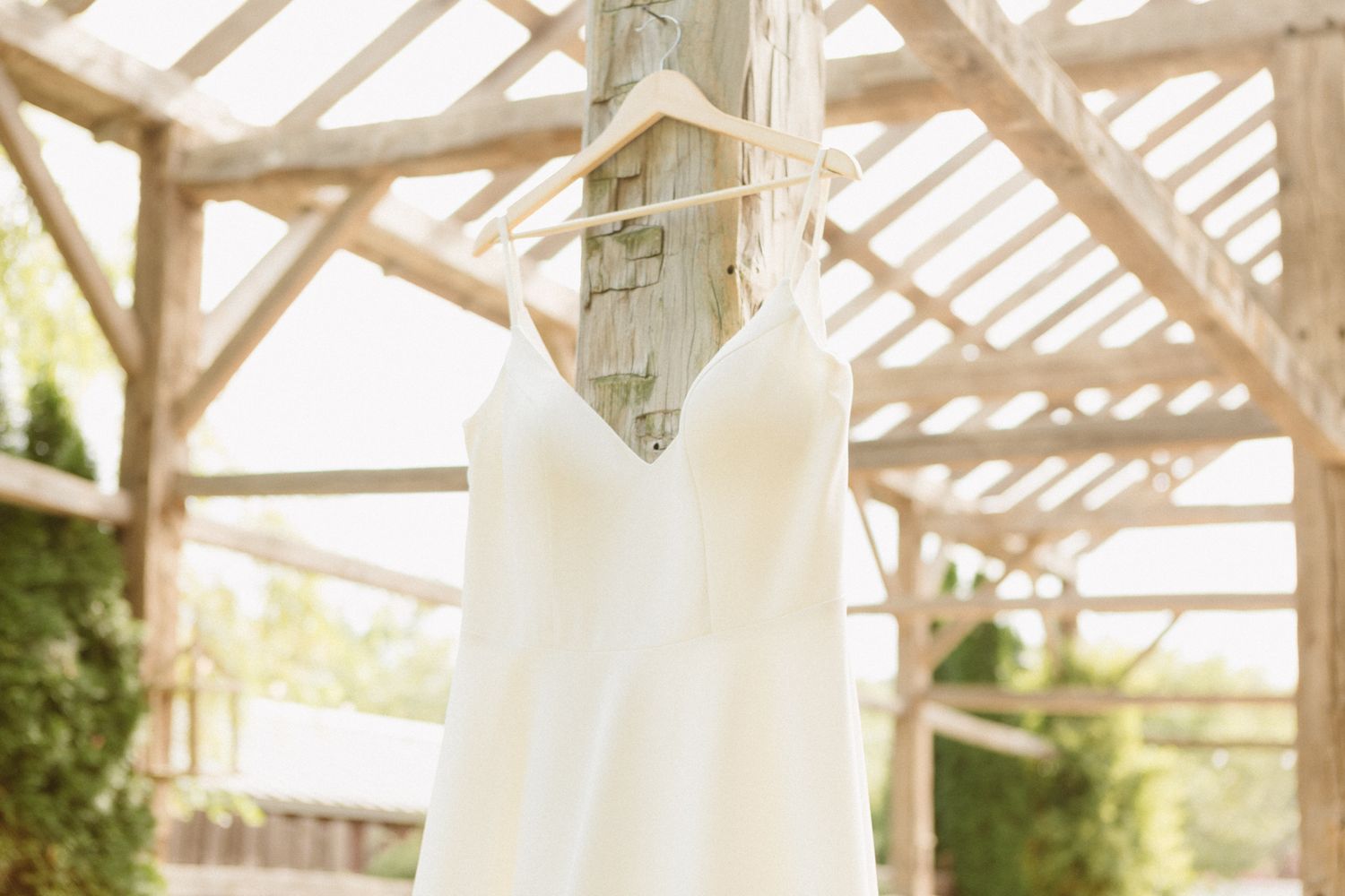 A white wedding dress hangs elegantly from a wooden pergola, backlit by soft natural sunlight filtering through the beams.
