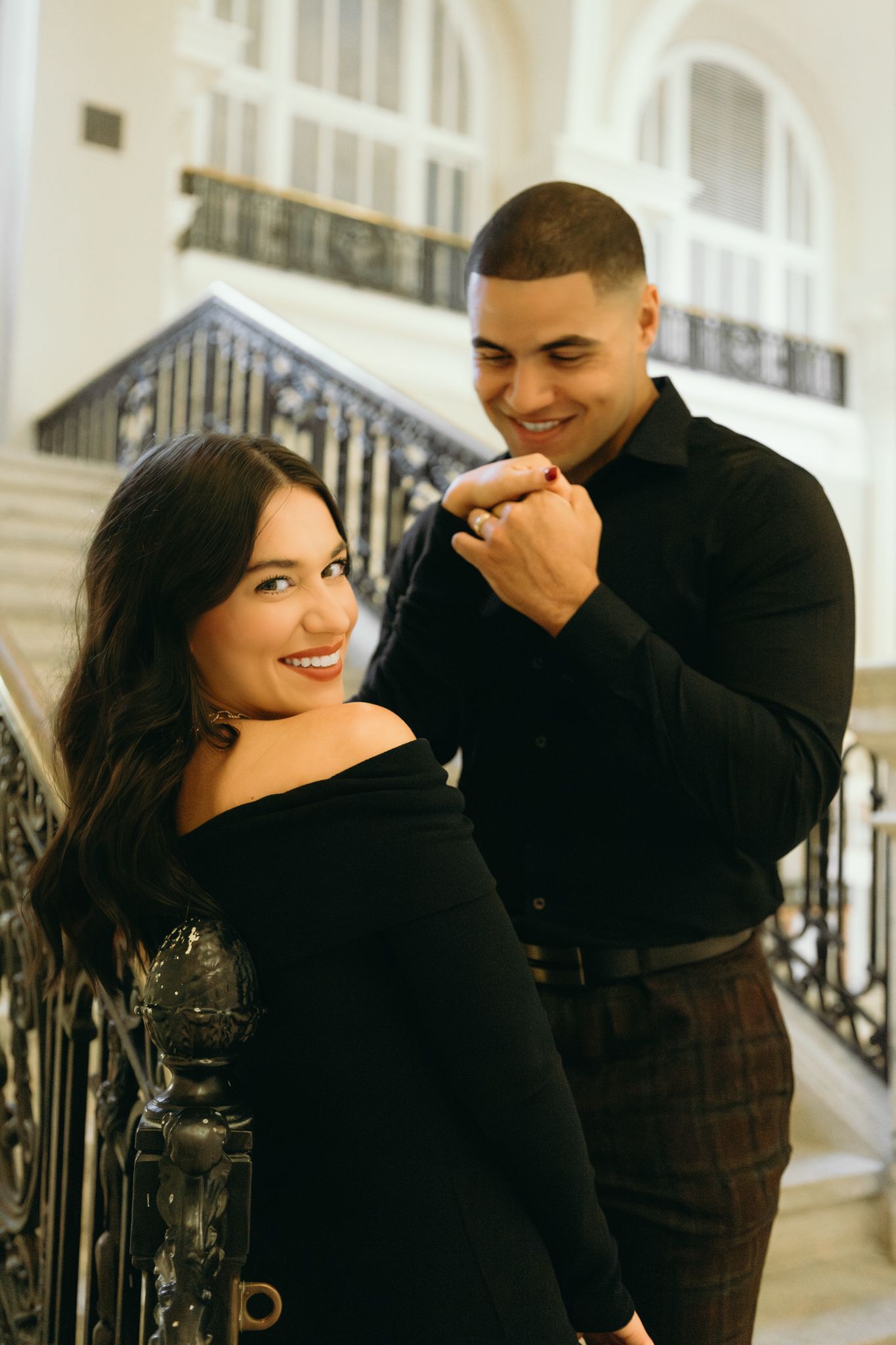 A couple poses elegantly on marble stairs indoors, both wearing stylish black outfits in a romantic setting.