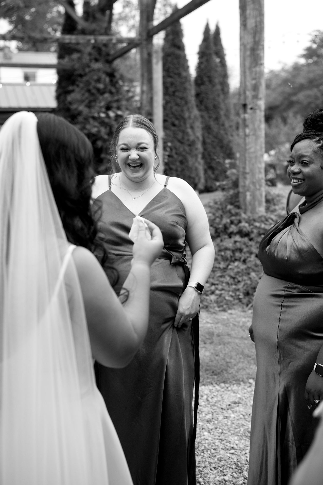 A black and white candid photo of bridesmaids sharing a joyful moment during a wedding in an outdoor garden setting.