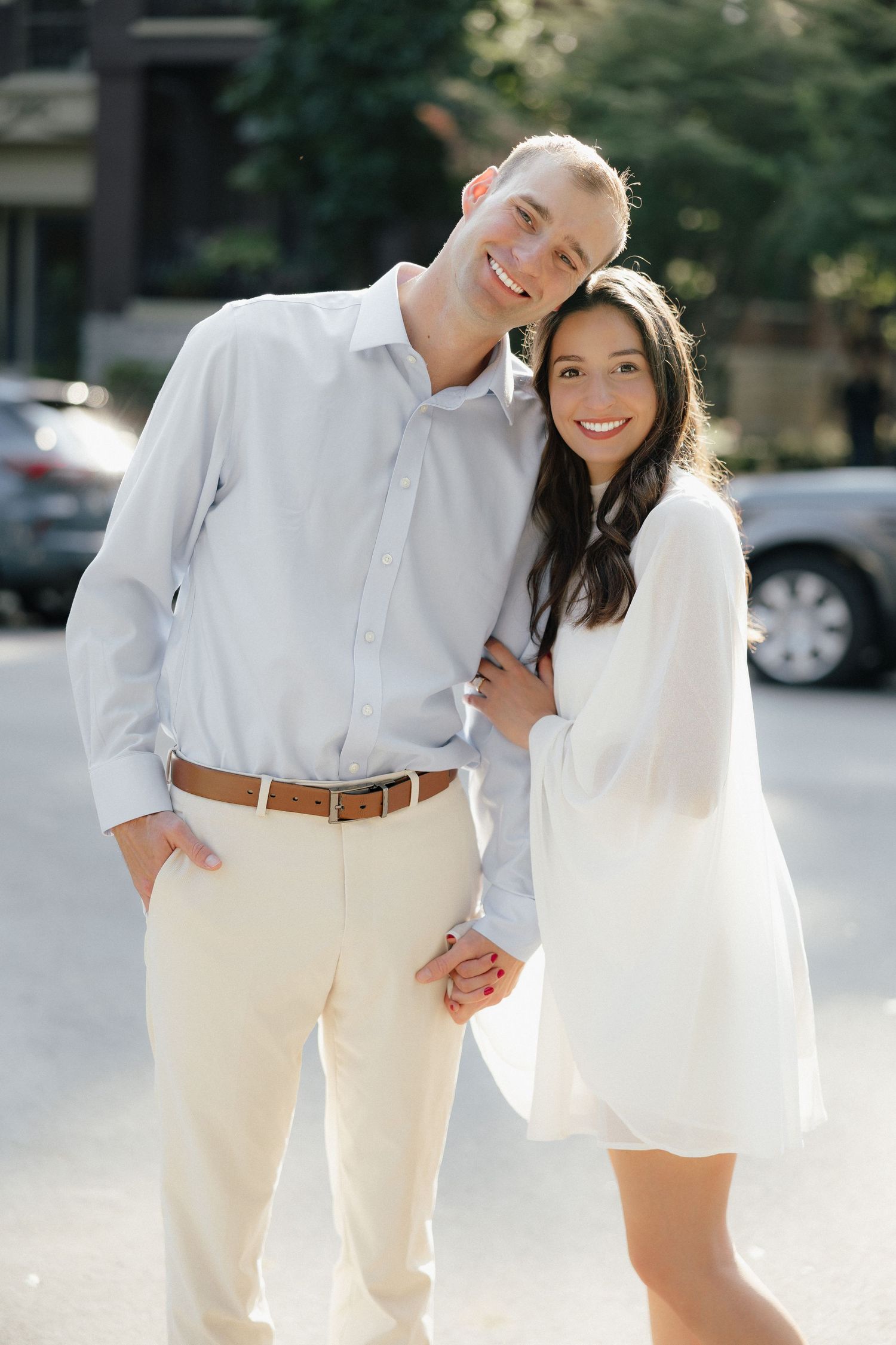 Smiling couple in white outfits holding hands on a sunny city street during an engagement photo session.
