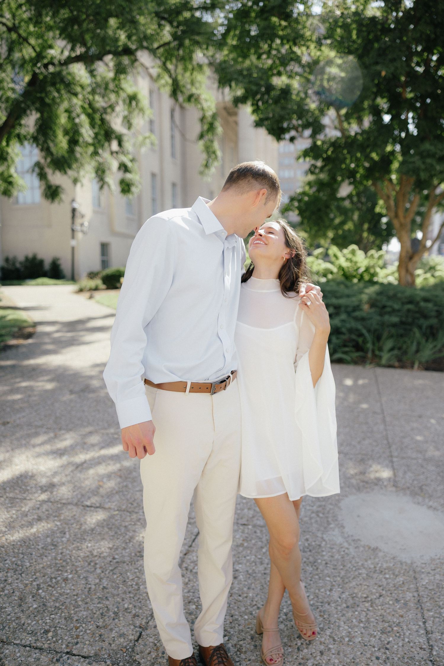 Couple in coordinating white and cream outfits sharing tender moment under shaded trees outdoors.
