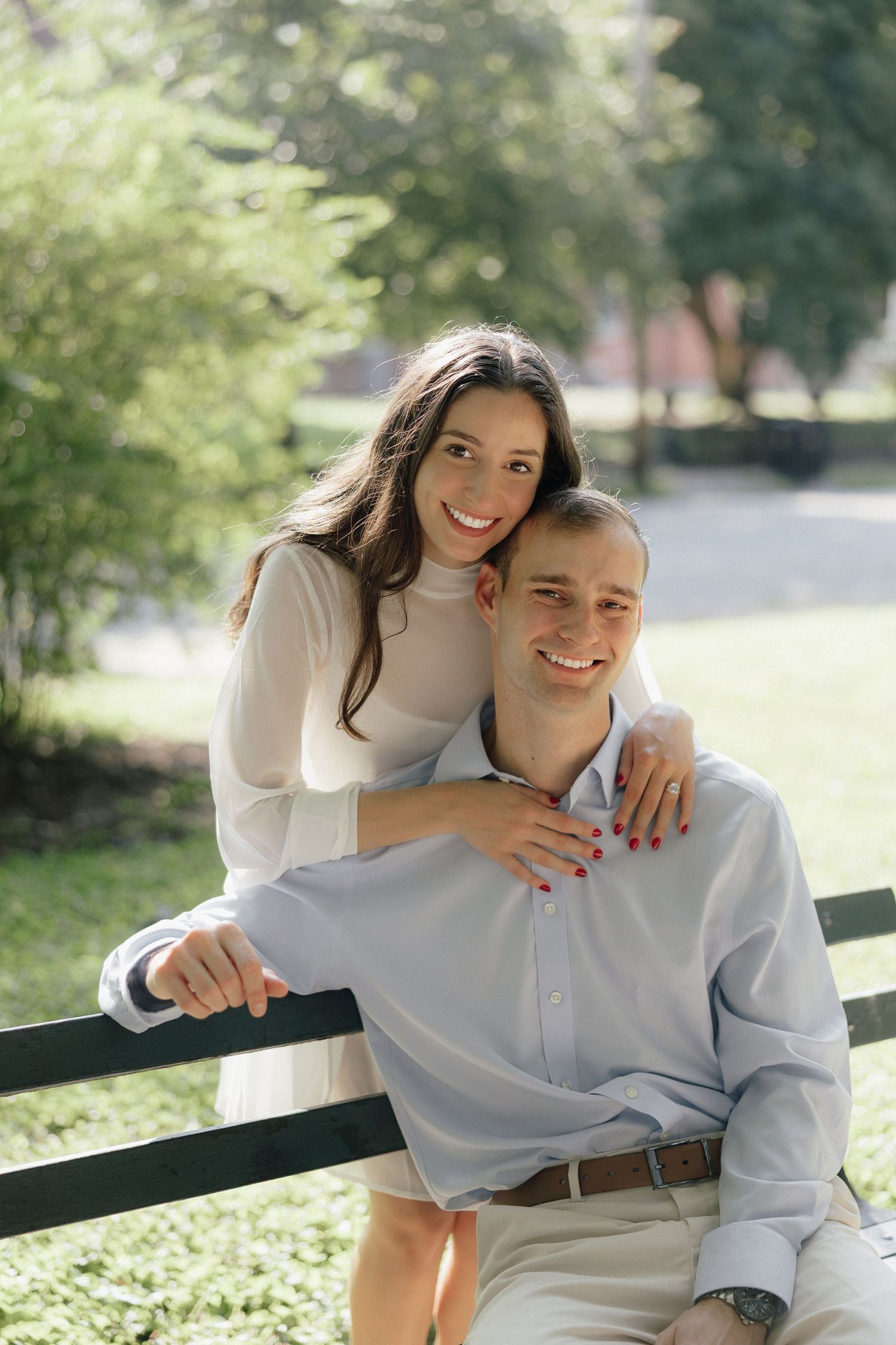 Smiling couple posing on a park bench, woman in white dress embracing man from behind outdoors.