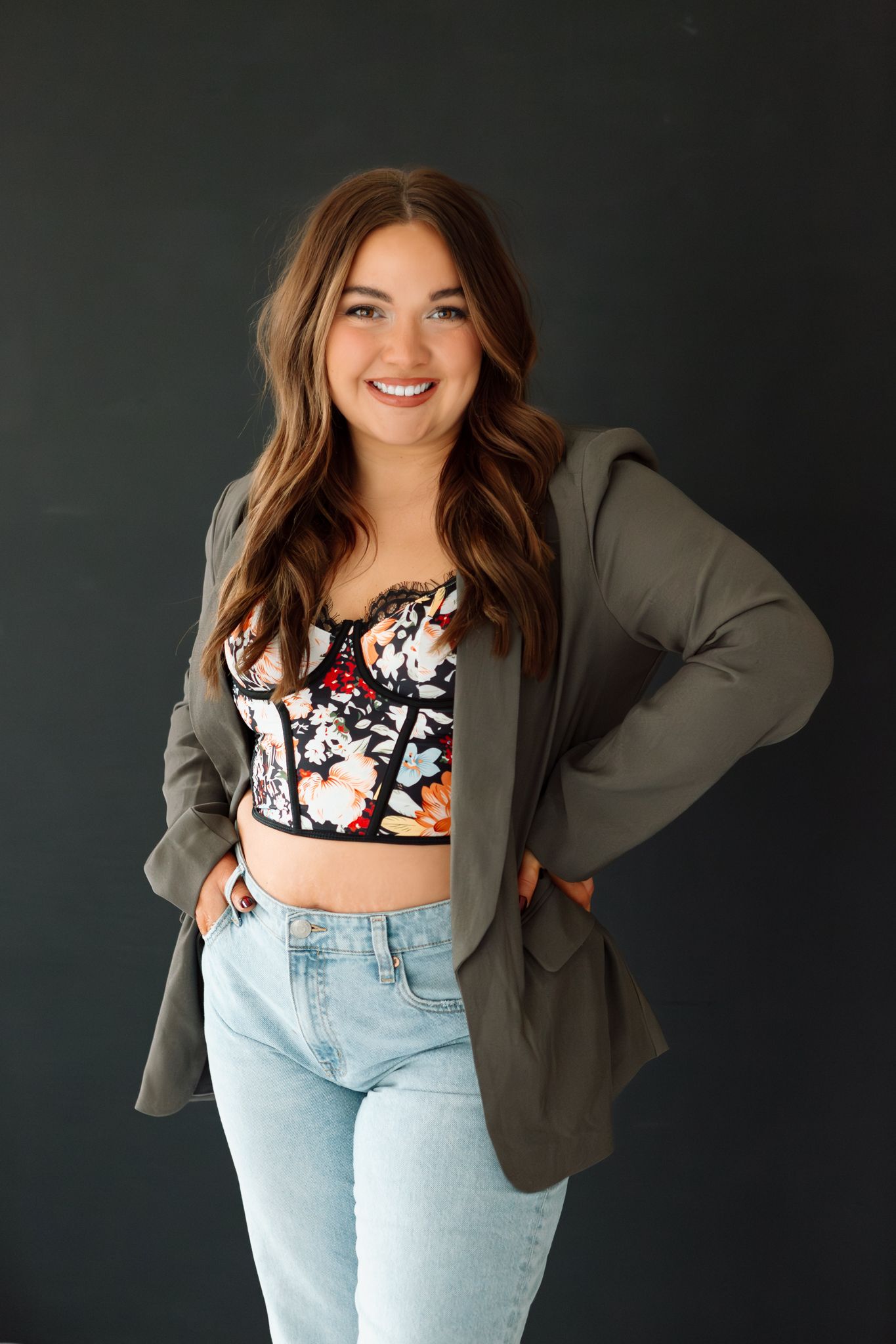 Smiling young woman in floral crop top, gray blazer, and light jeans posing against dark background.