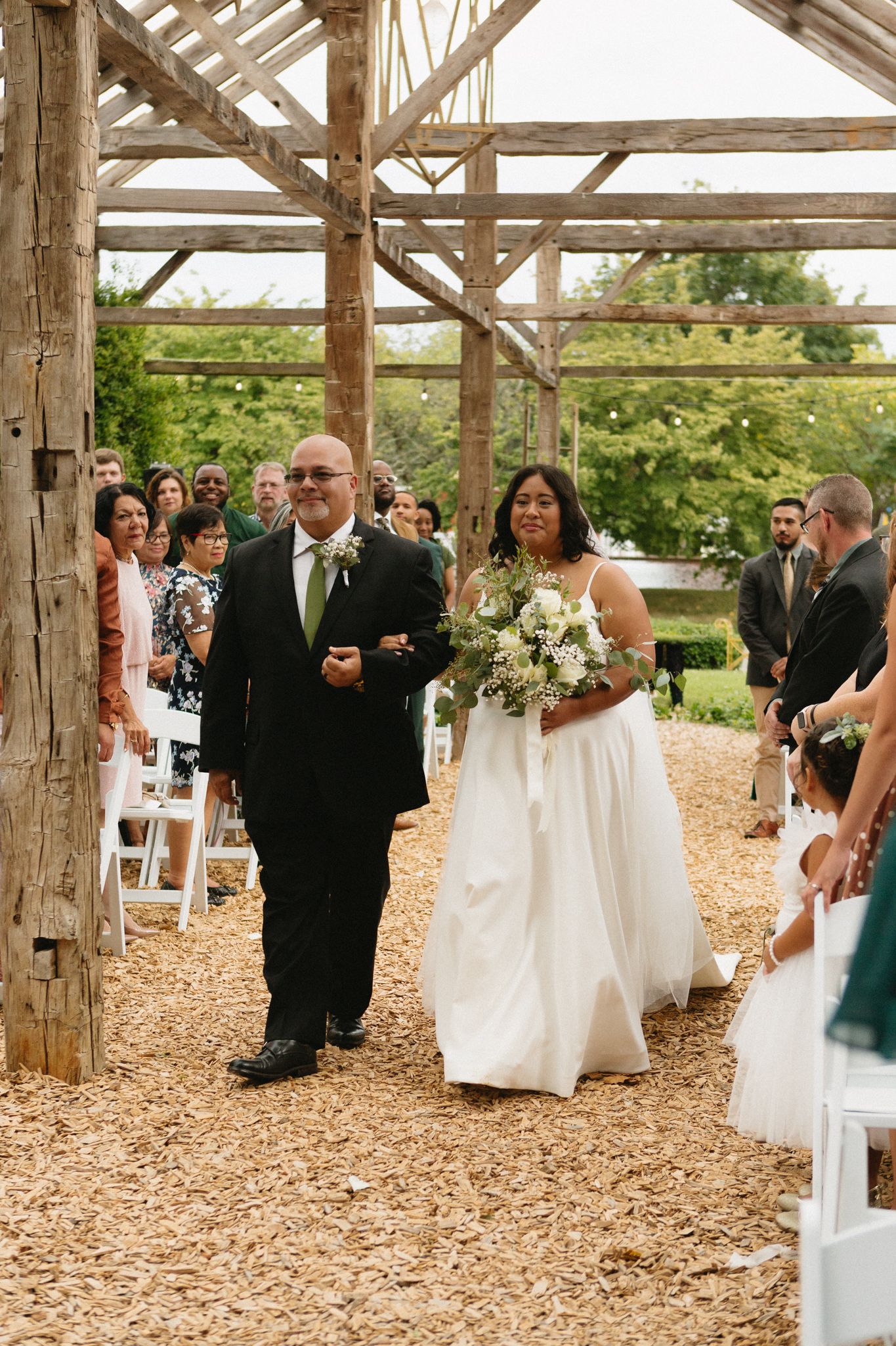 A wedding ceremony takes place in a rustic wooden barn decorated with white chairs along a mulch-covered aisle.