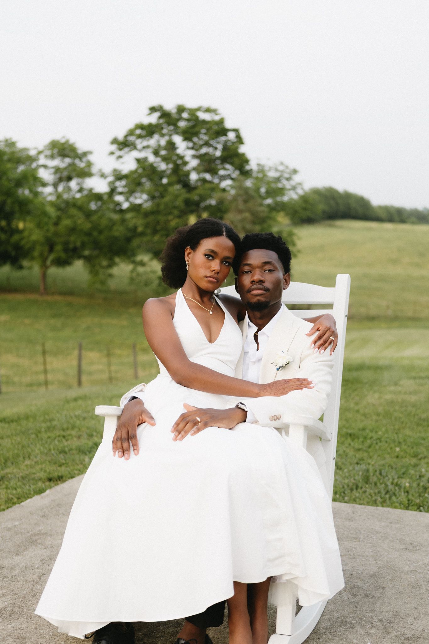 Couple dressed in white pose together on rustic chair in outdoor countryside setting.