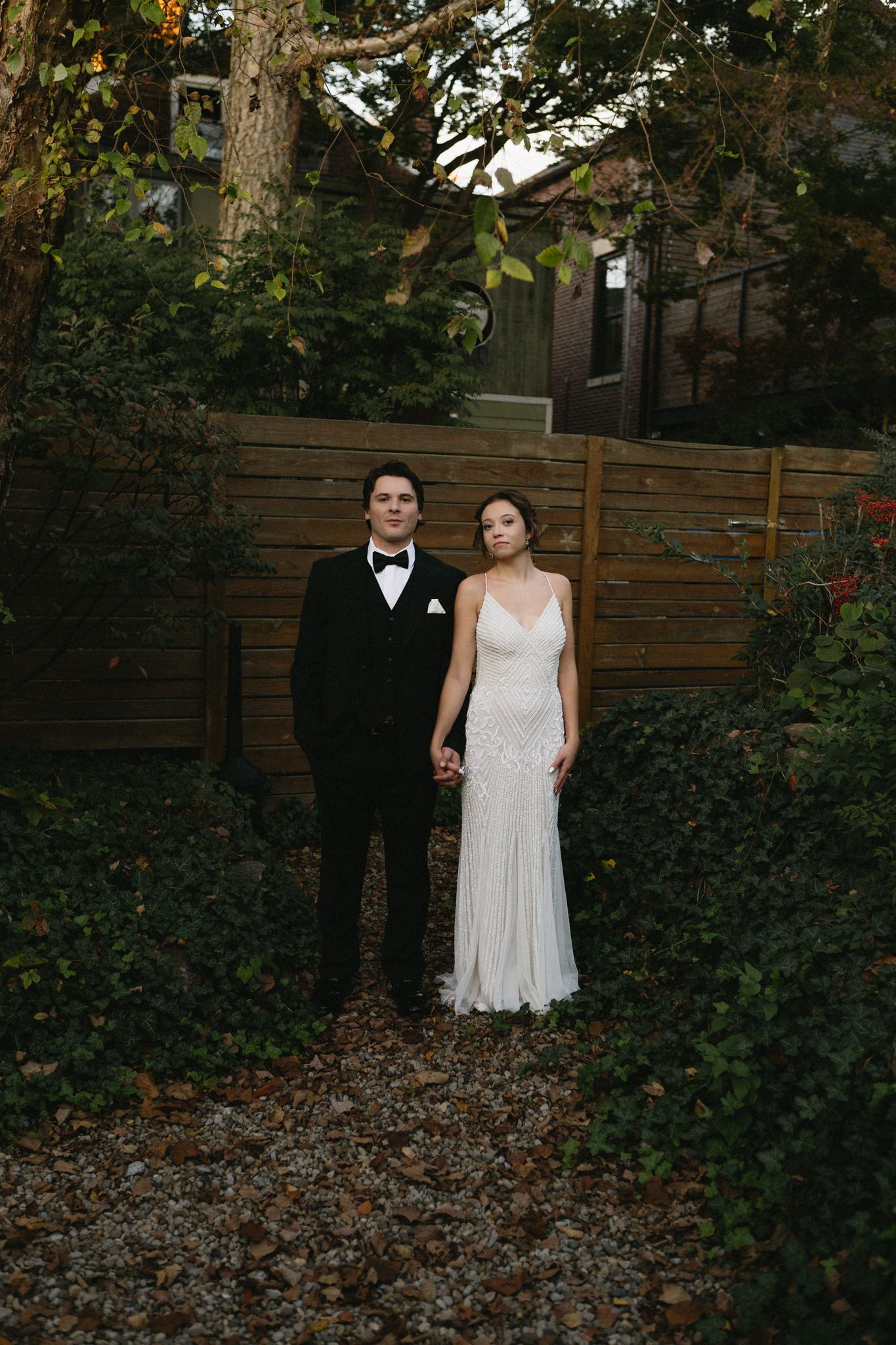 A wedding couple poses in front of a wooden fence surrounded by lush greenery.