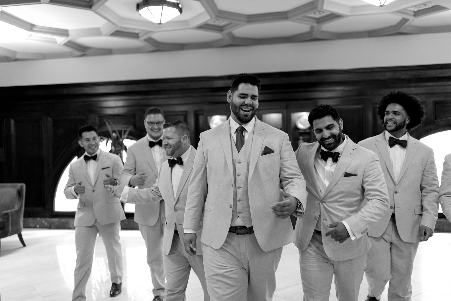 Black and white photo showing a wedding party in light colored suits walking and smiling indoors.