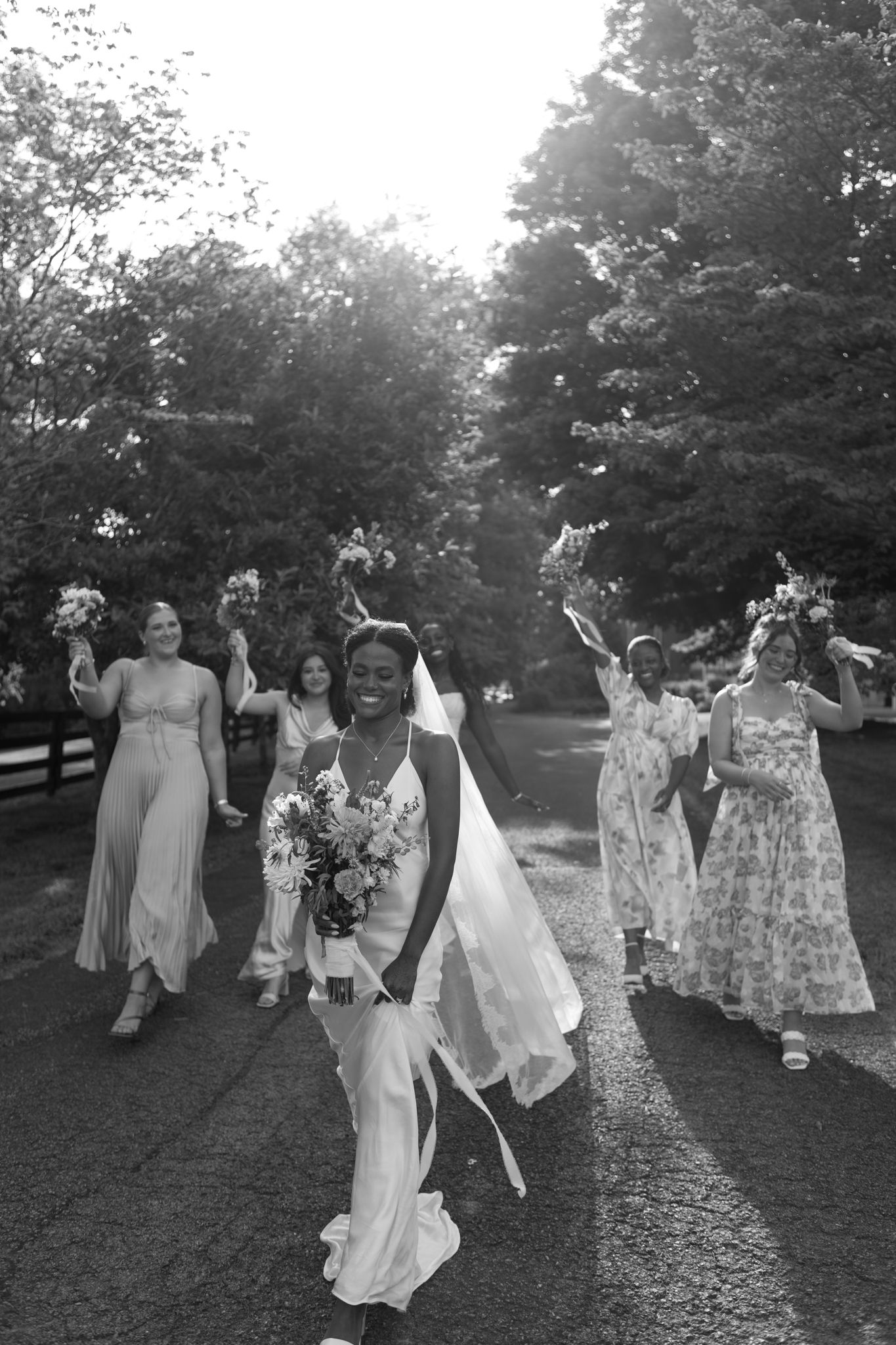 Wedding party celebrates joyfully on a grassy lawn during golden hour in black and white photography.