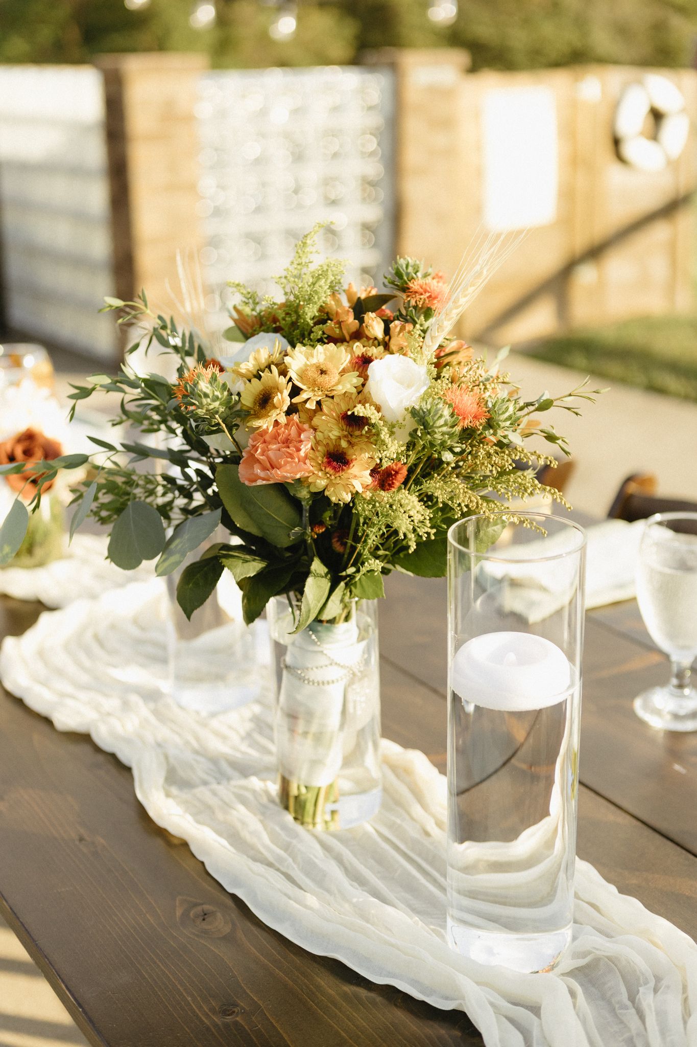Elegant floral centerpieces with white fabric runners decorate a rustic wooden table at a wedding reception.
