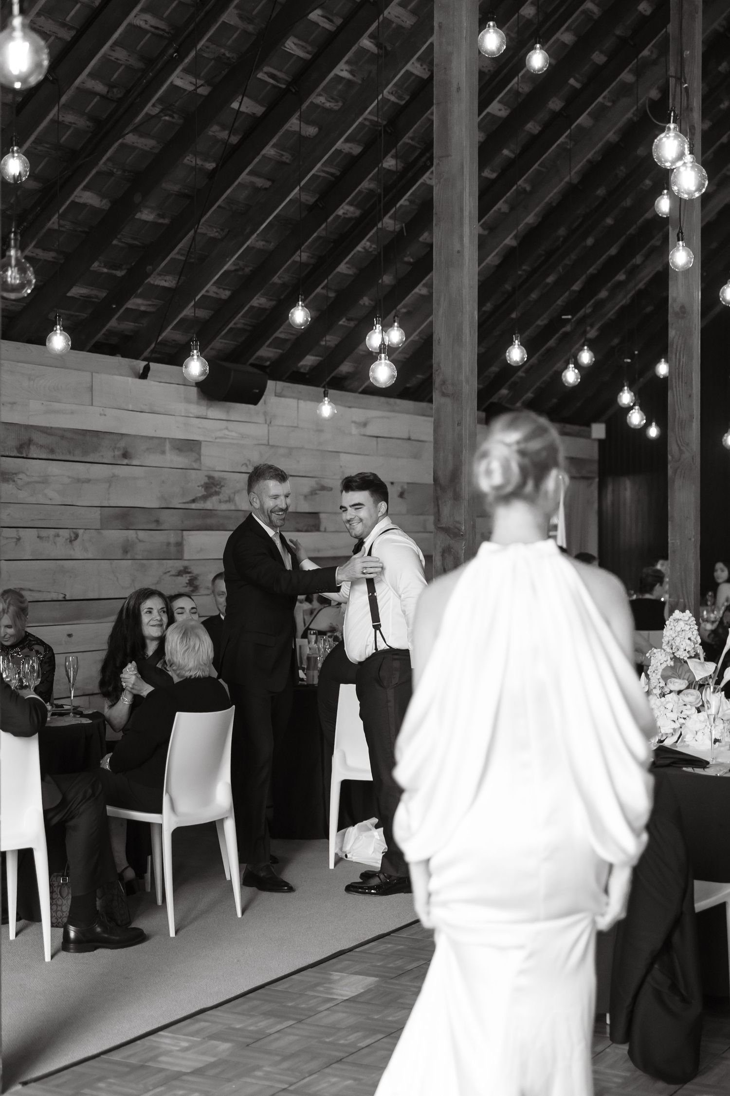 Groom shaking hands with groomsman at elegant barn wedding reception with string lights.