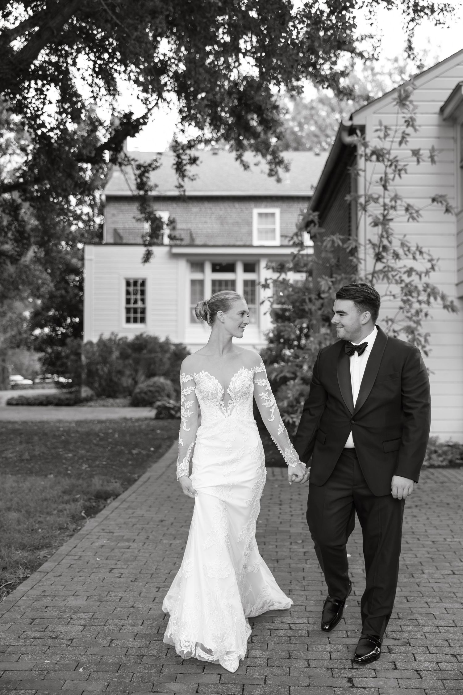 Bride and groom walk hand-in-hand along a brick path in black and white photo outside a historic estate.