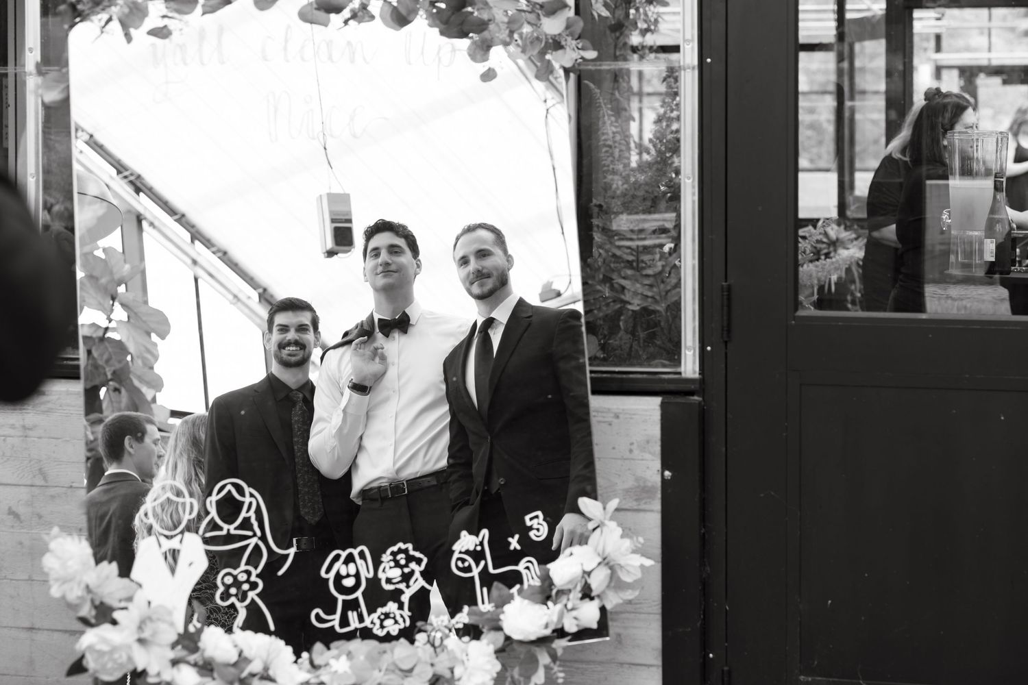 Three men in tuxedos posing at wedding reception with decorative cake toppers in black and white photo.