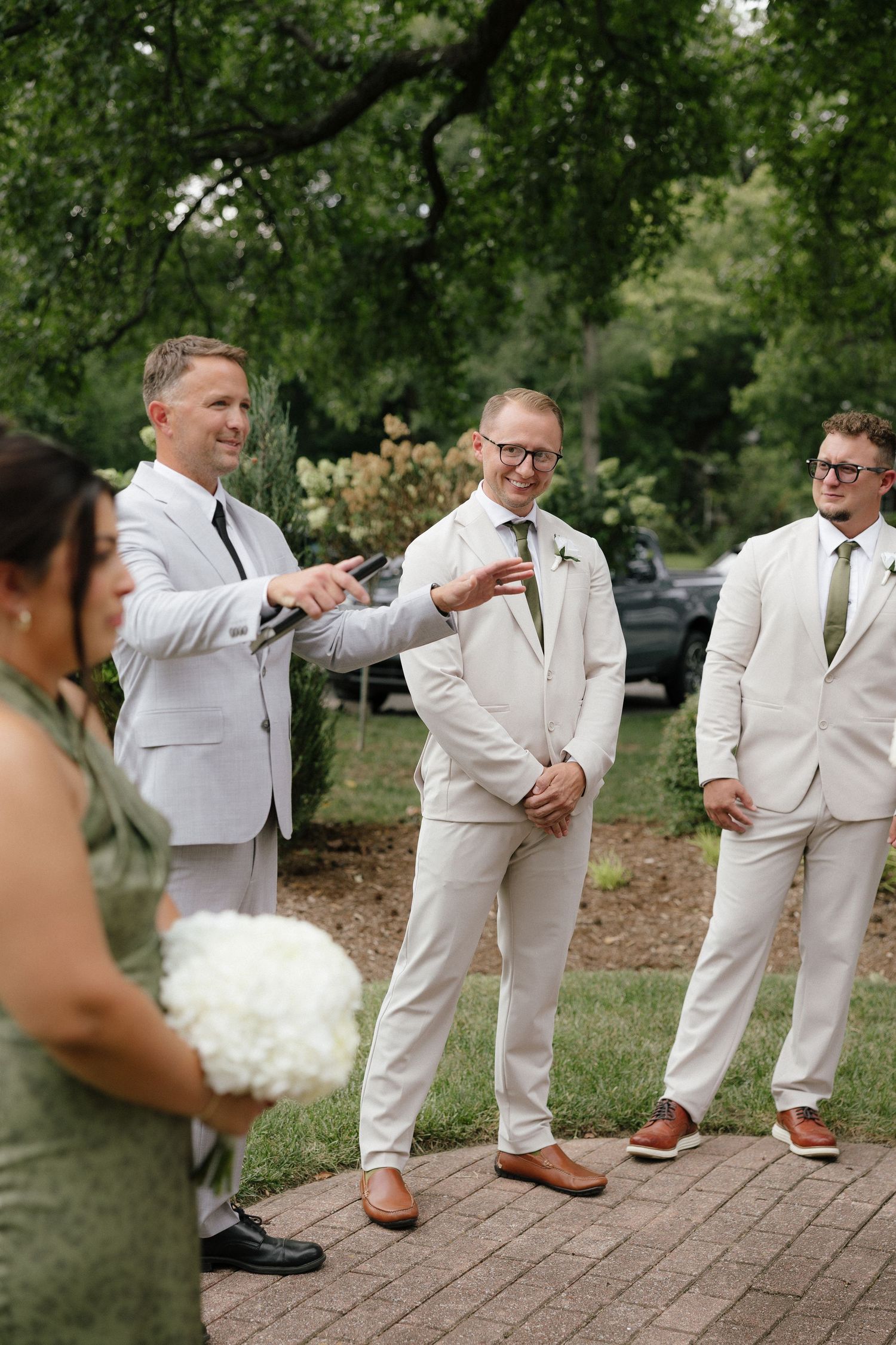 Groom smiles with groomsmen in light suits on brick pathway during outdoor wedding ceremony.