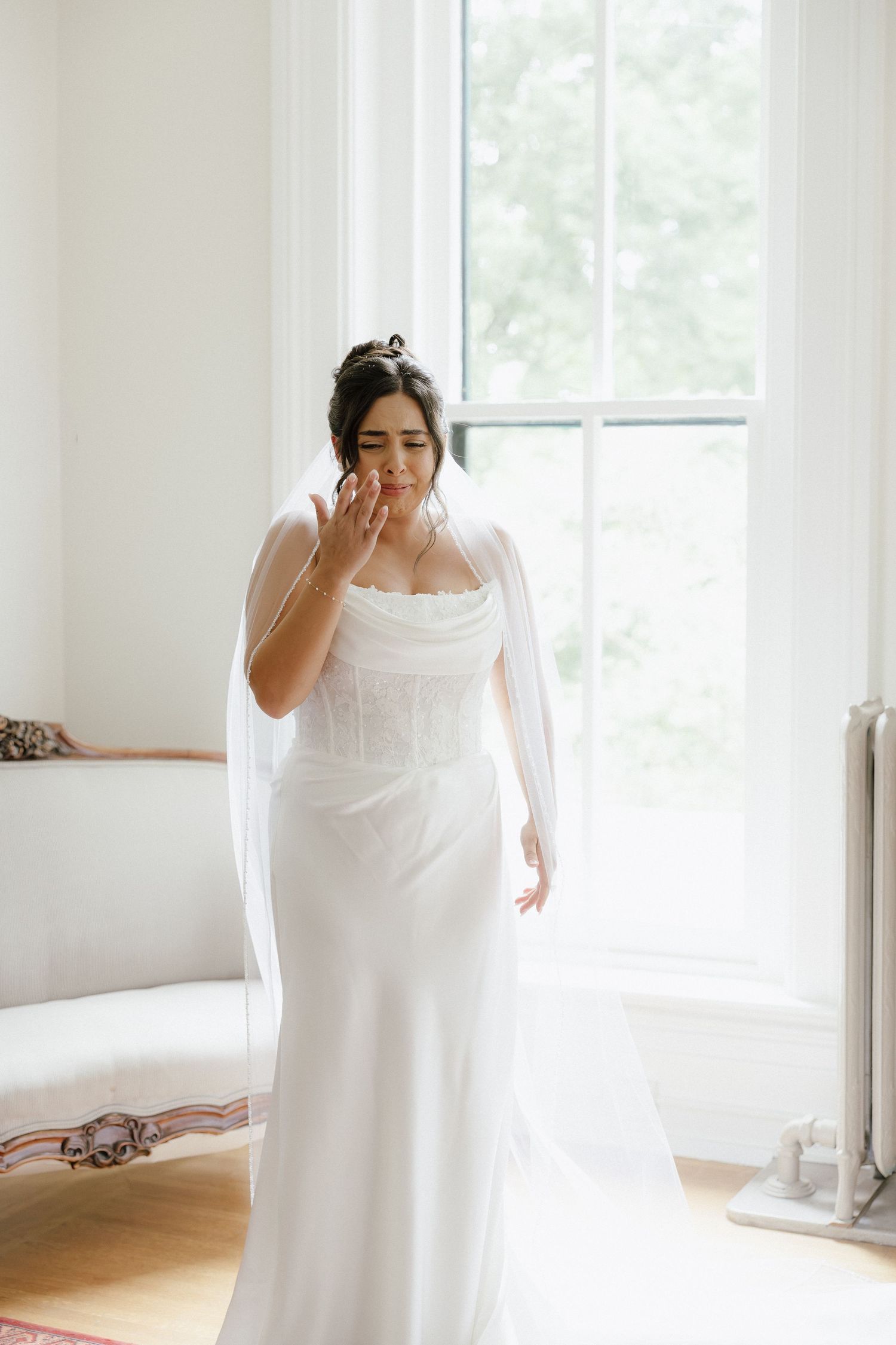 Emotional bride in a fitted white corset wedding gown wiping tears by a bright window before ceremony.