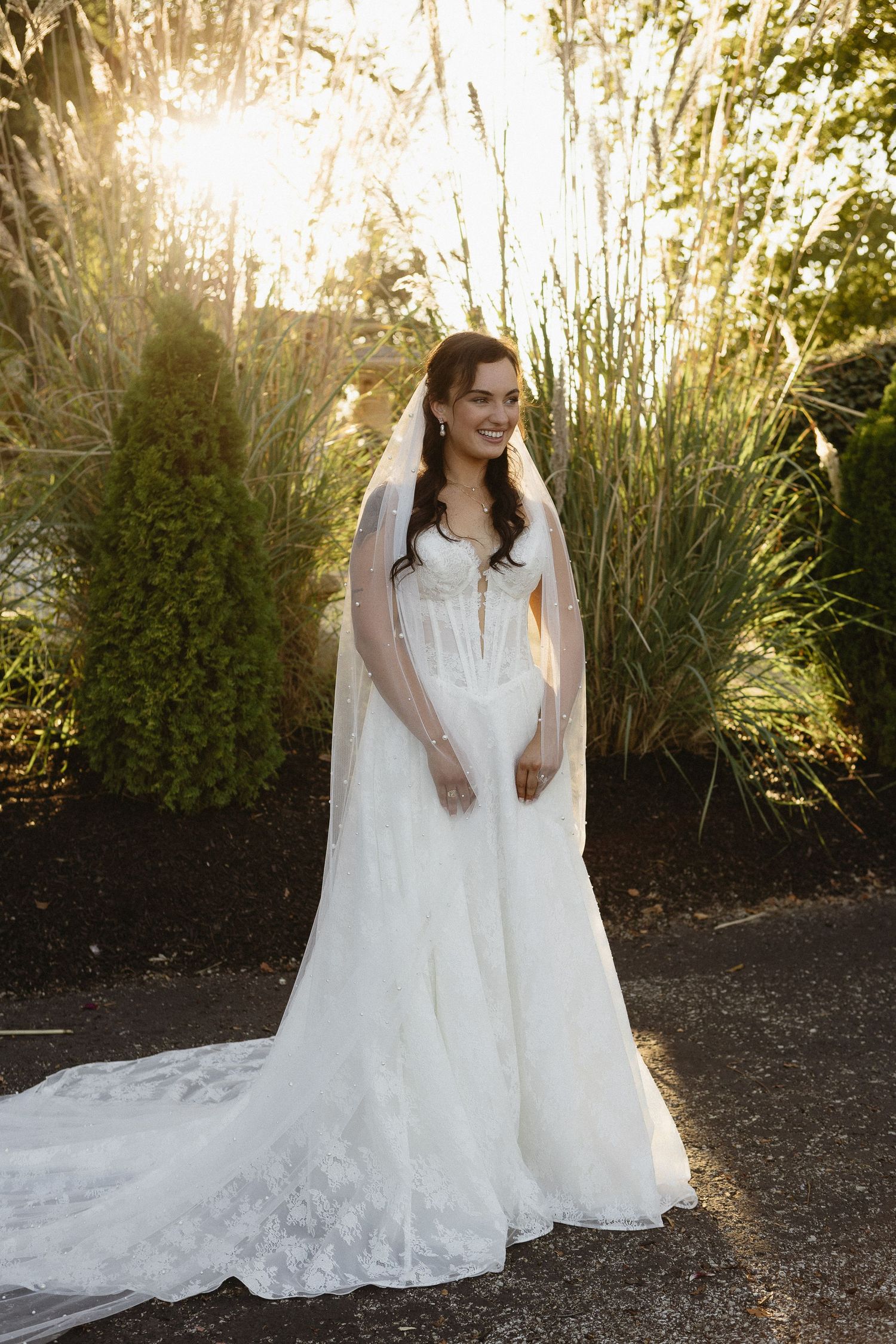 White wedding dresses with flowing trains photographed in golden sunlight against garden foliage.