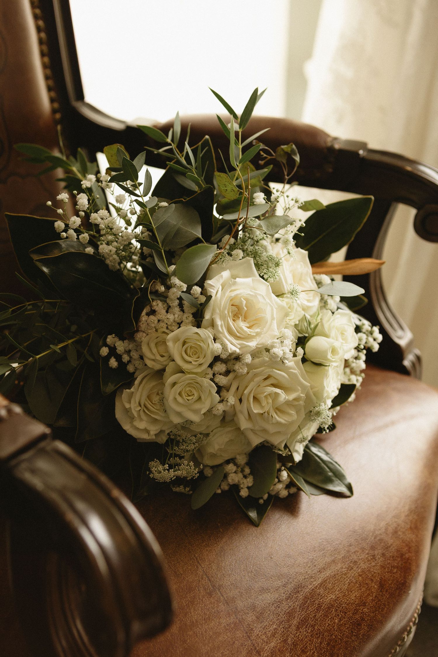 A classic white rose wedding bouquet with baby's breath and greenery rests on a brown leather chair.
