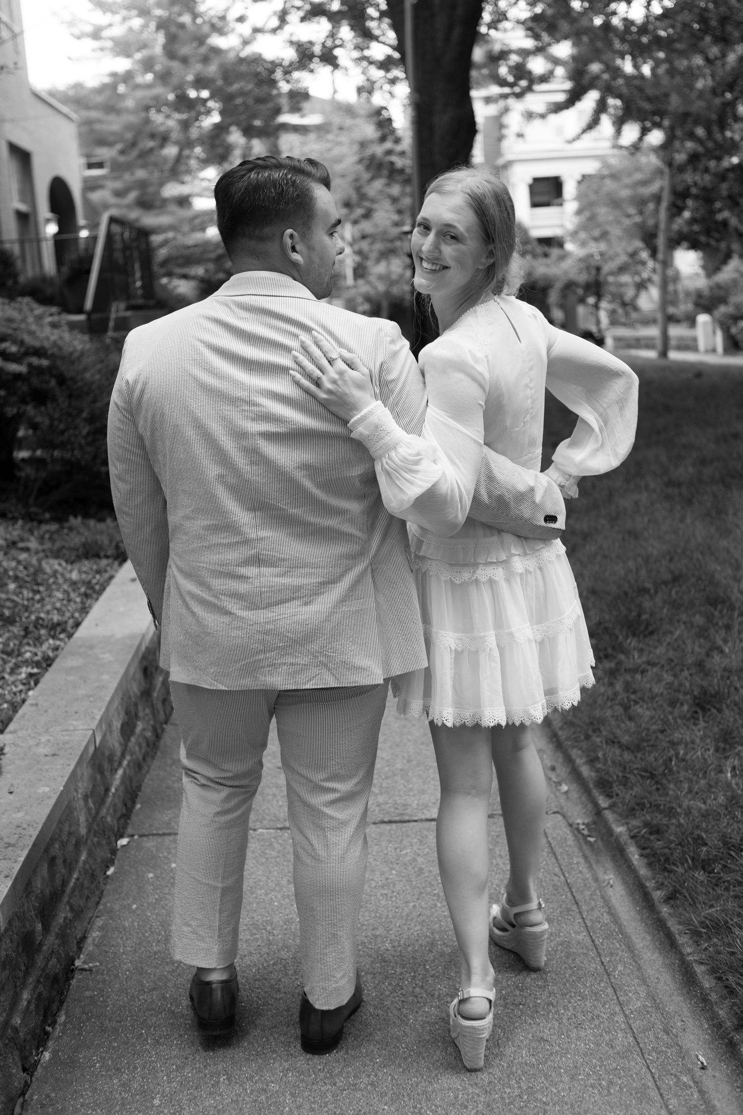 Black and white photo of a couple walking on a sidewalk, woman in white dress smiling at man in light suit.