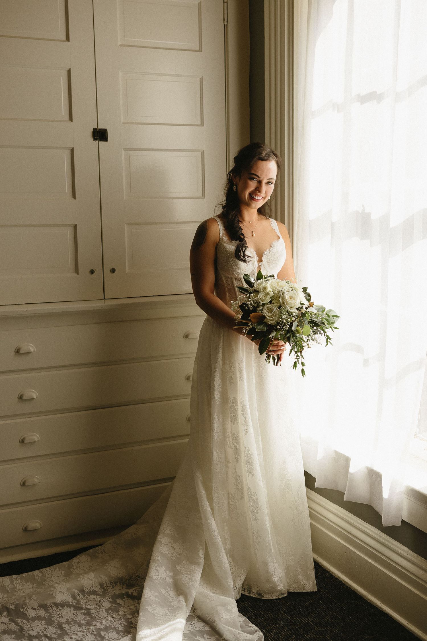 A bride in a flowing white dress poses near a window in a room with white built-in cabinets.