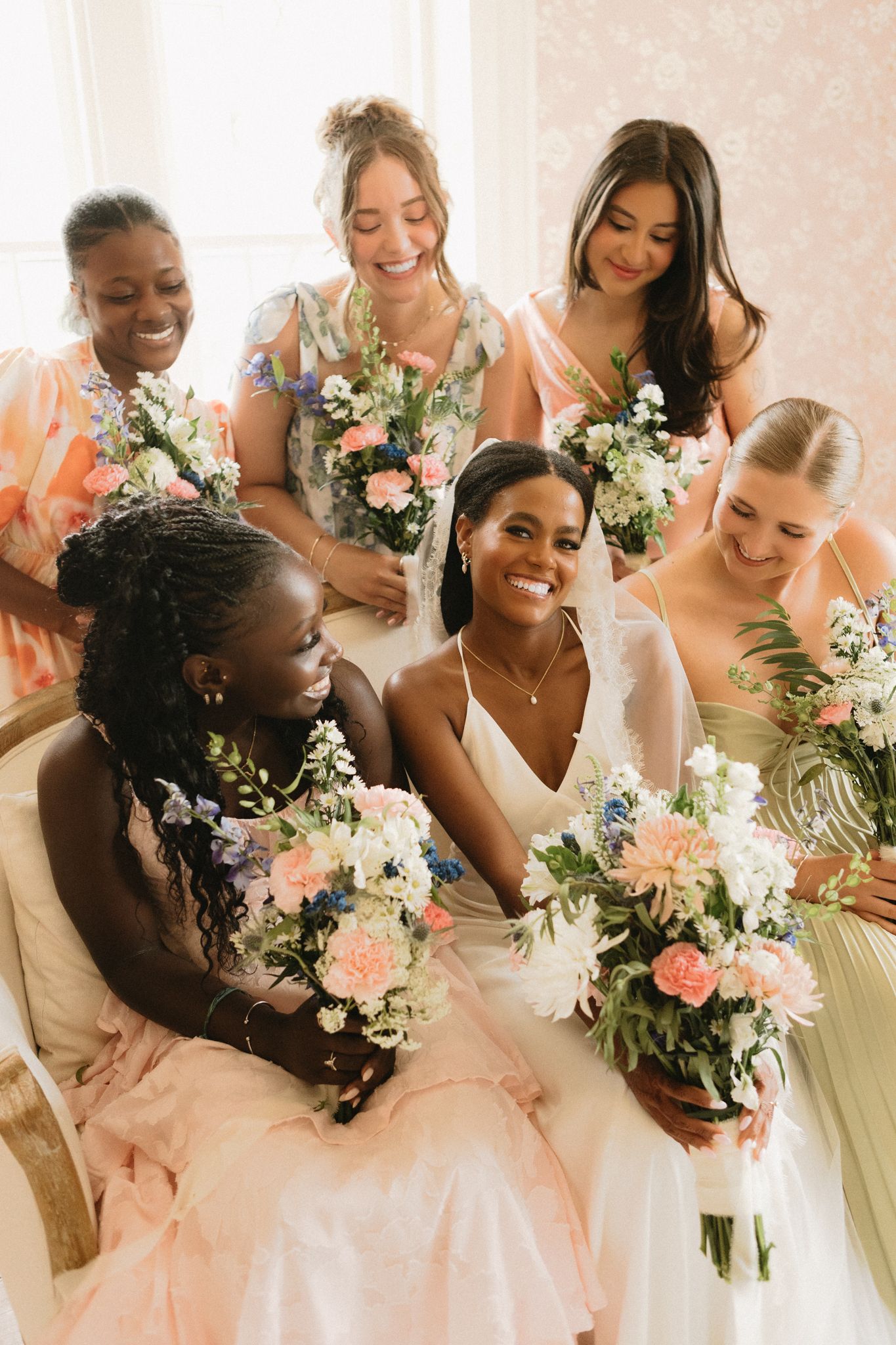 Bridesmaids in pastel dresses hold beautiful pink and white flower bouquets while posing together.