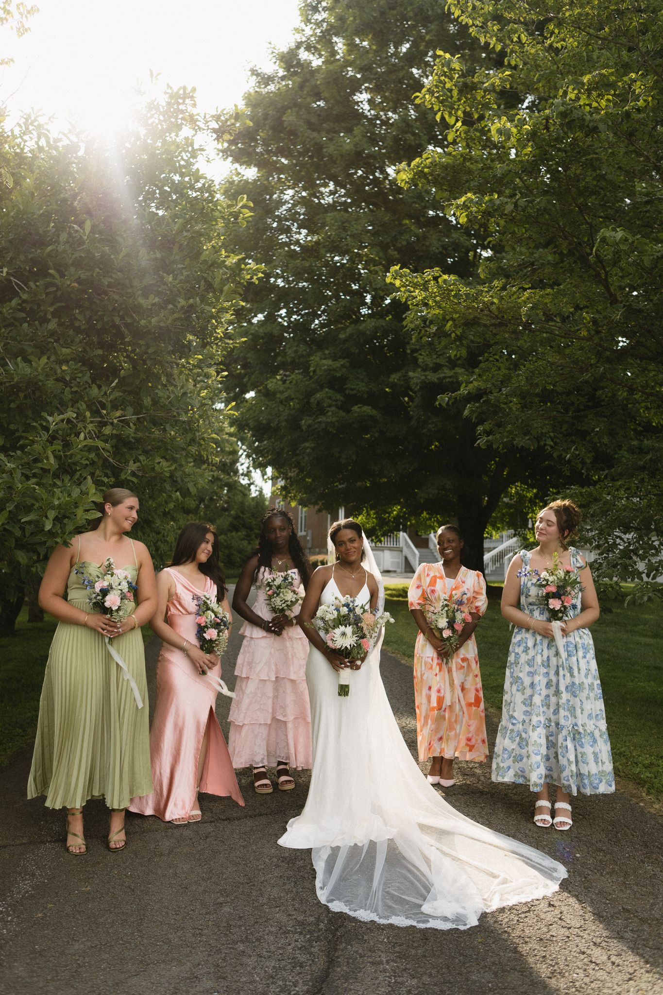 A bridal party in pastel floral dresses poses on a tree-lined path during golden hour.