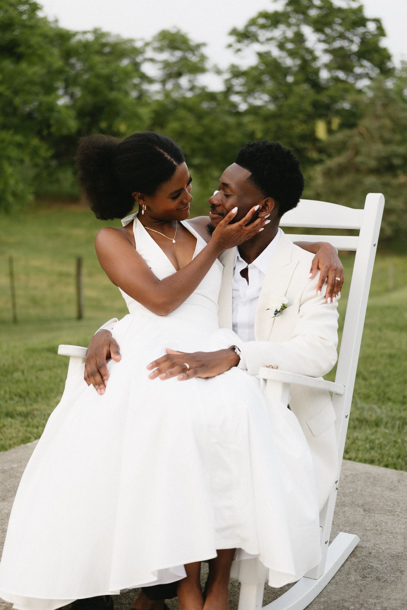 Two people in white outfits share a romantic moment together outdoors on rustic wooden chairs.