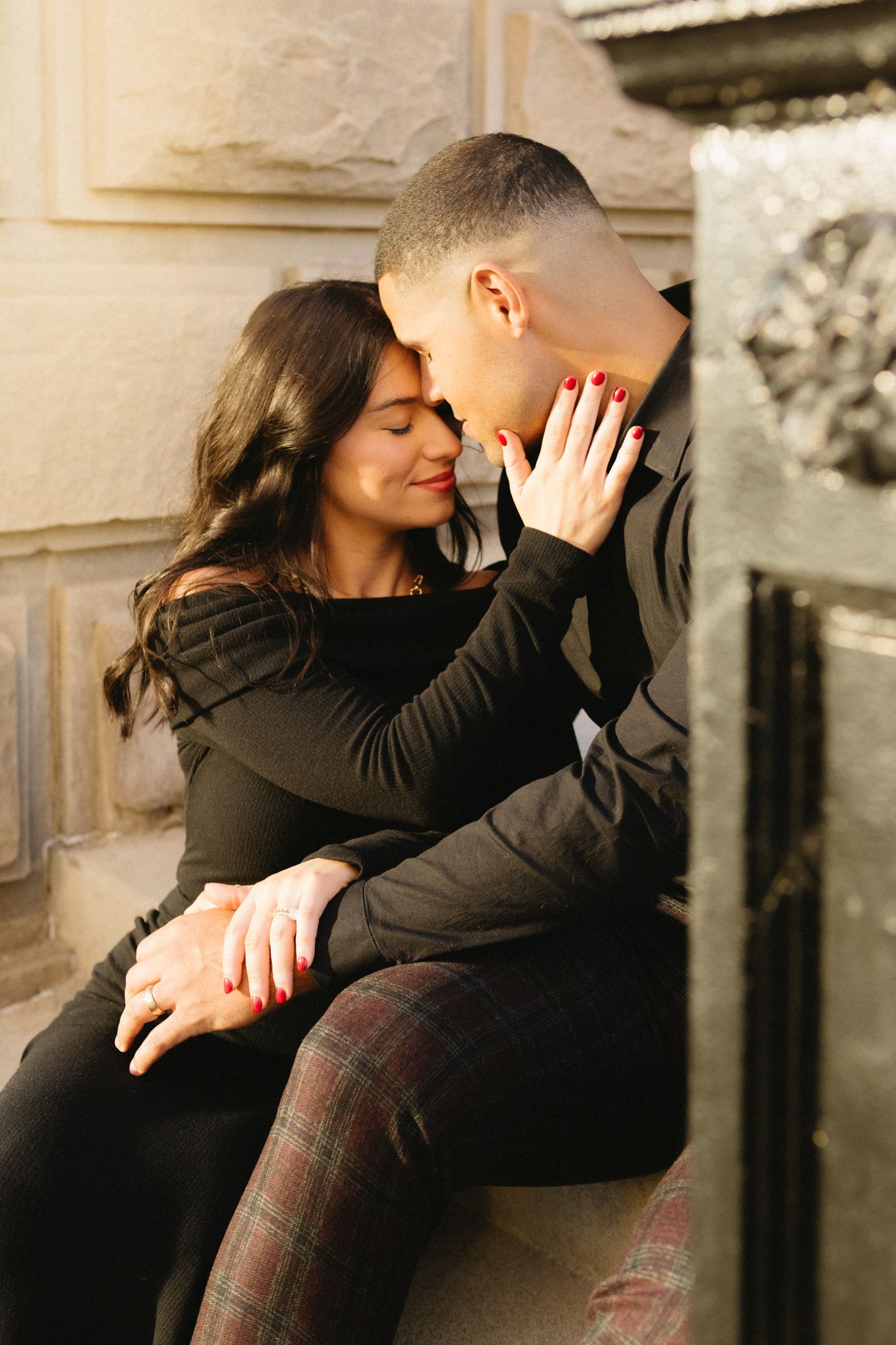 Couple in dark clothing sharing an affectionate moment while sitting together outdoors near stone architecture.