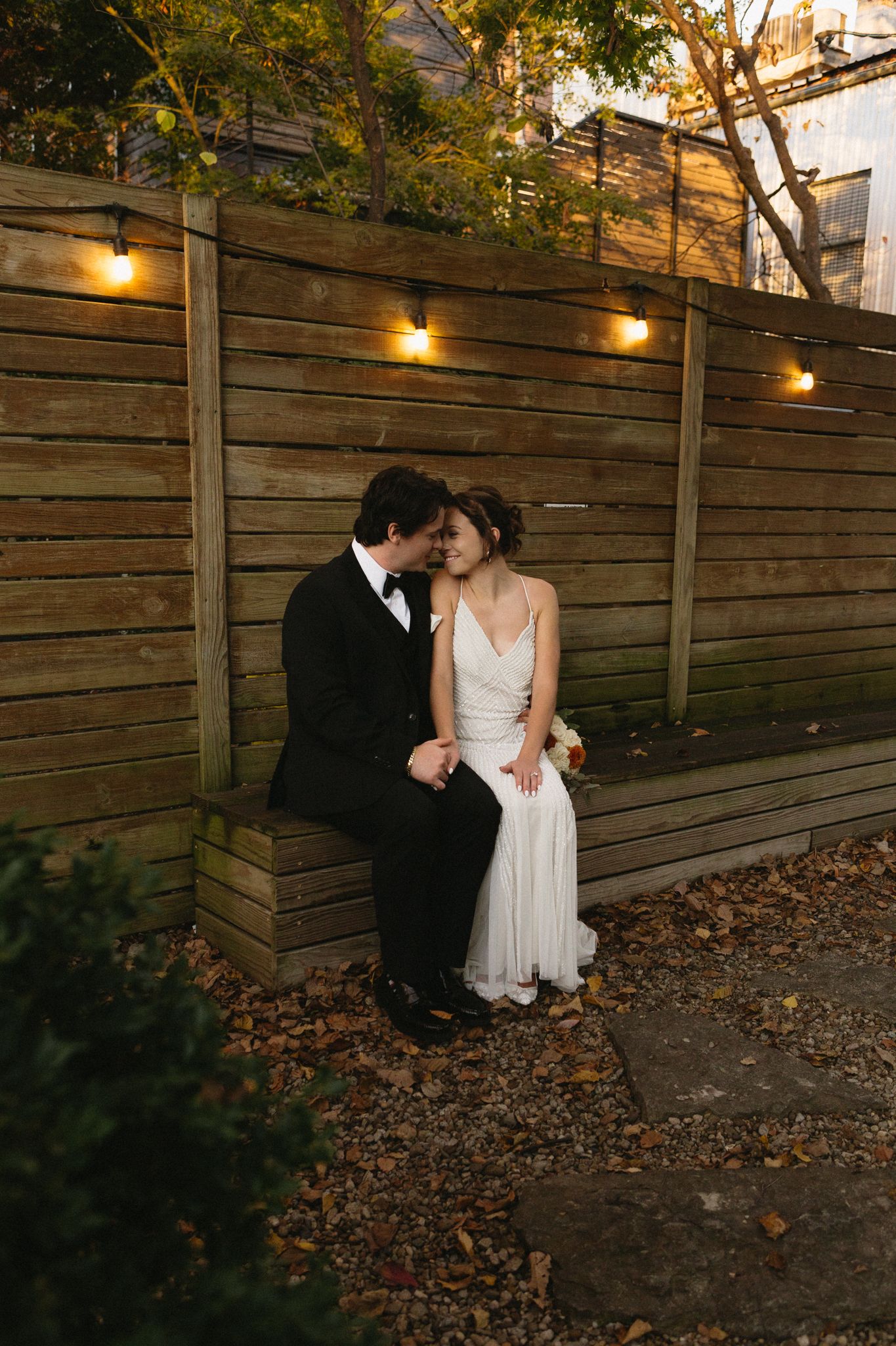 Wedding couple shares an intimate moment against a wooden wall with string lights at dusk.