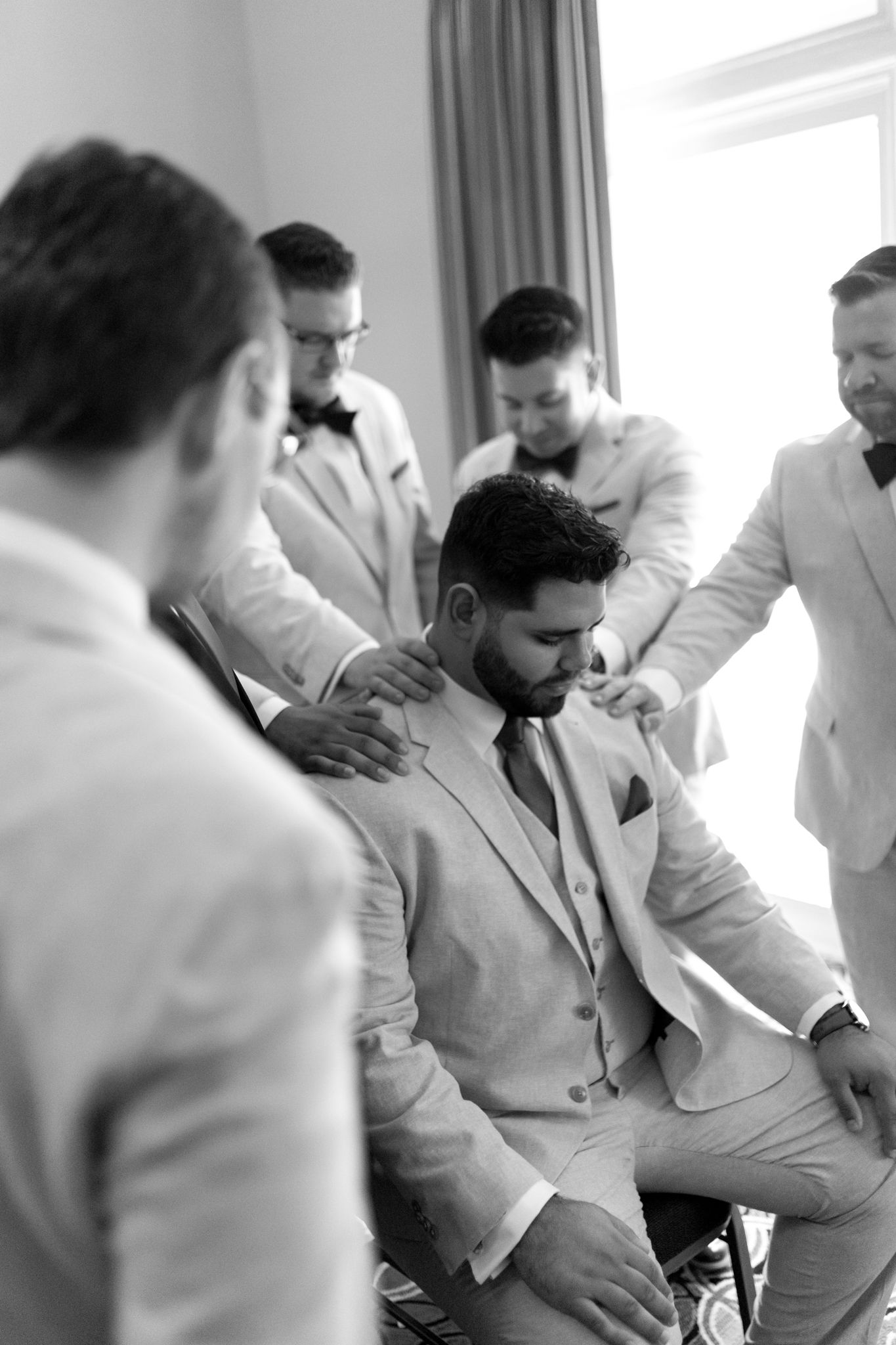 A group of groomsmen in light colored suits gather around during pre-wedding preparations in a sunlit room.