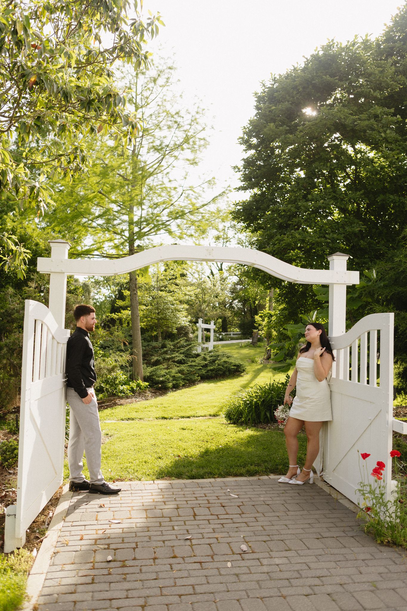 A couple poses along a stone pathway with white garden gates and lush greenery in the background.