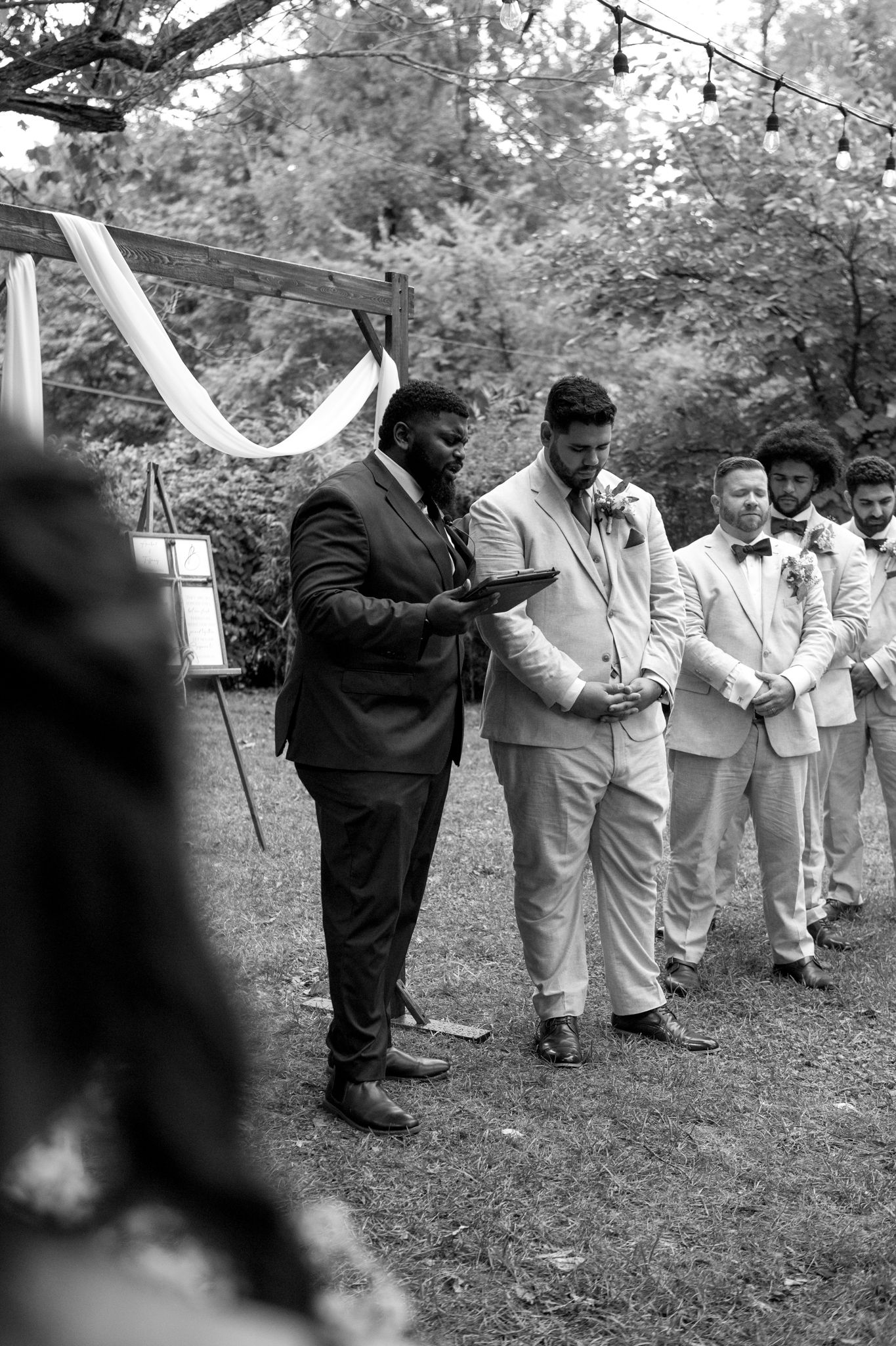 Black and white wedding photo showing groomsmen gathered outside with string lights and white fabric decor overhead.
