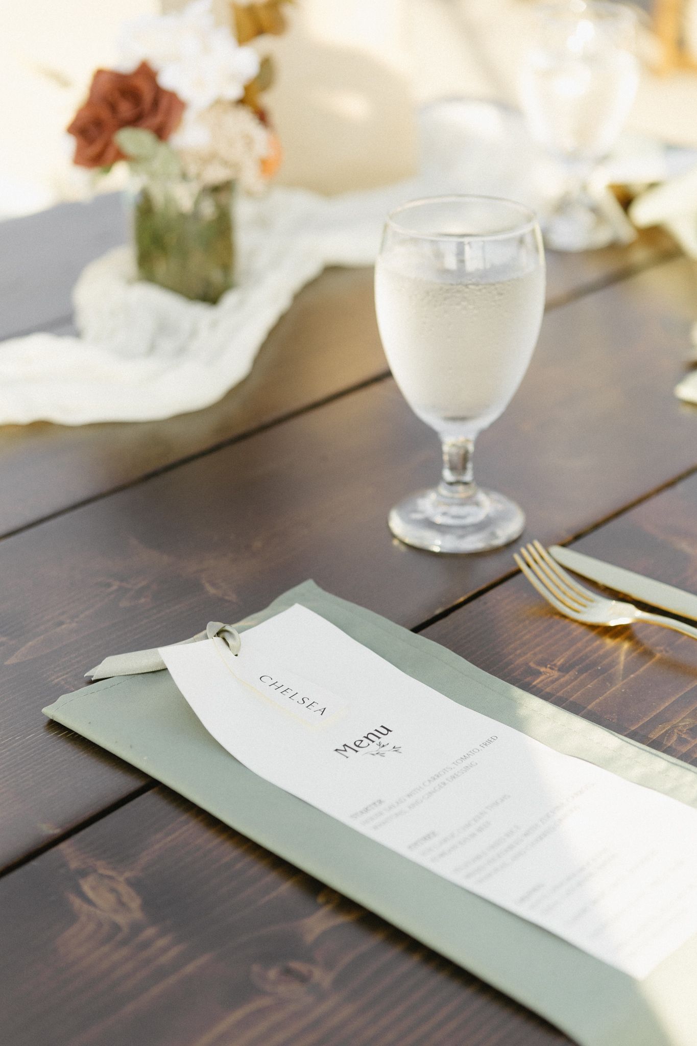 Wedding reception table setting with elegant menu cards in sage green and white glassware on dark wood.