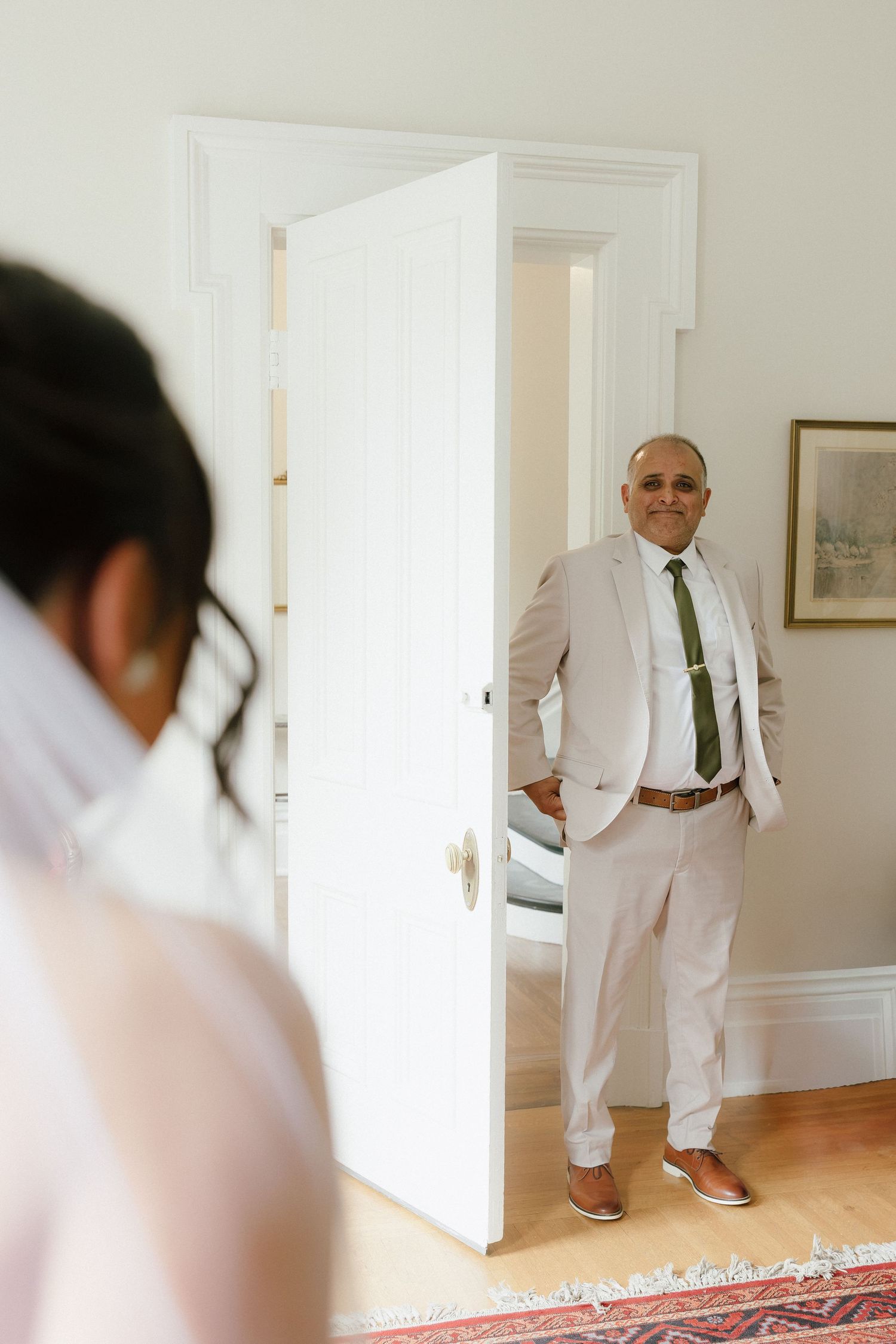Father of the bride smiling in a beige suit and green tie, standing in a doorway during wedding preparations.