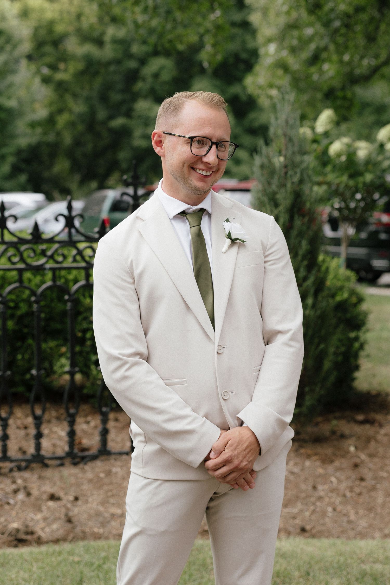 Smiling groom in cream suit with green tie and boutonniere standing outdoors.