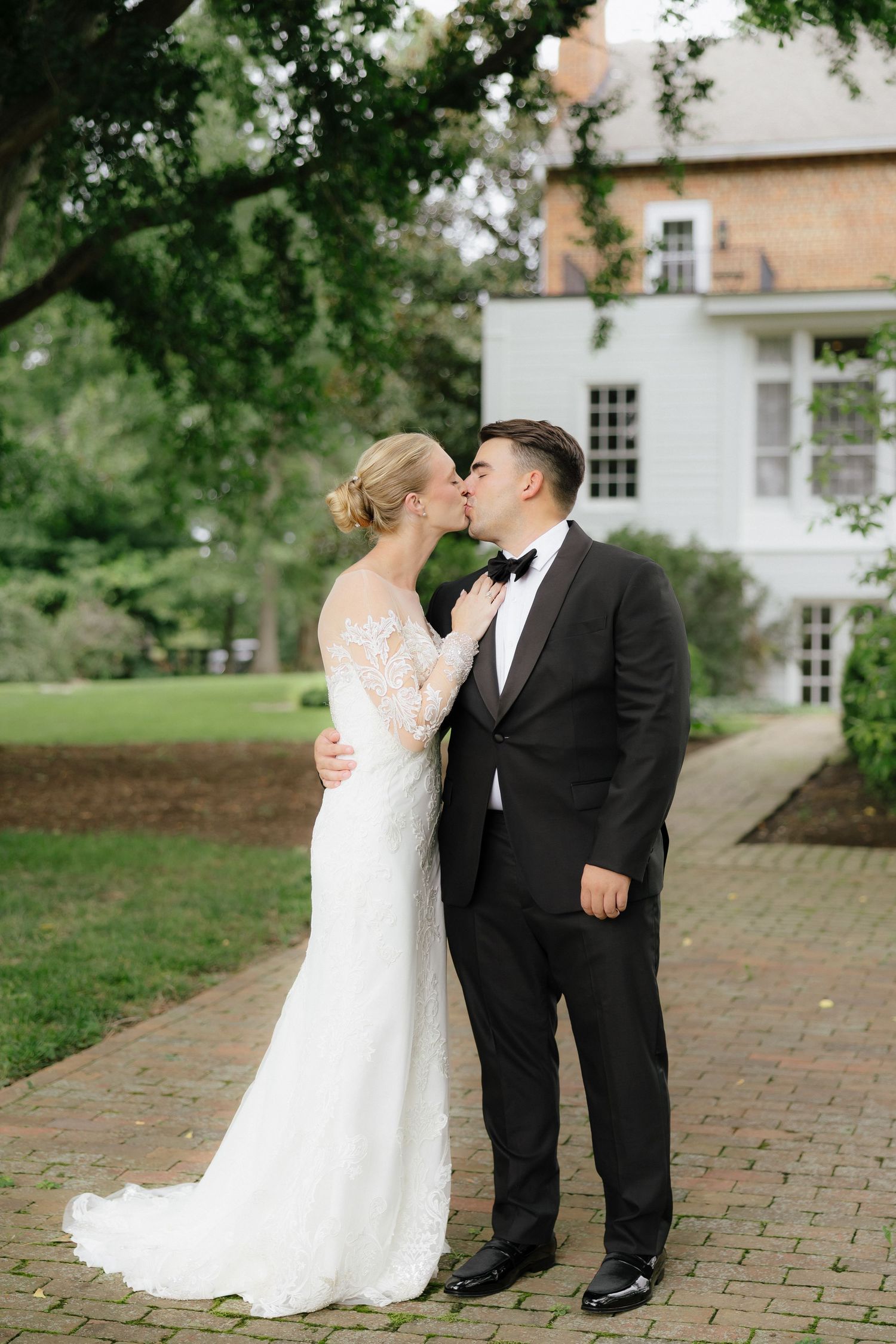 Bride and groom sharing a kiss outdoors on brick path with elegant white manor house in background.