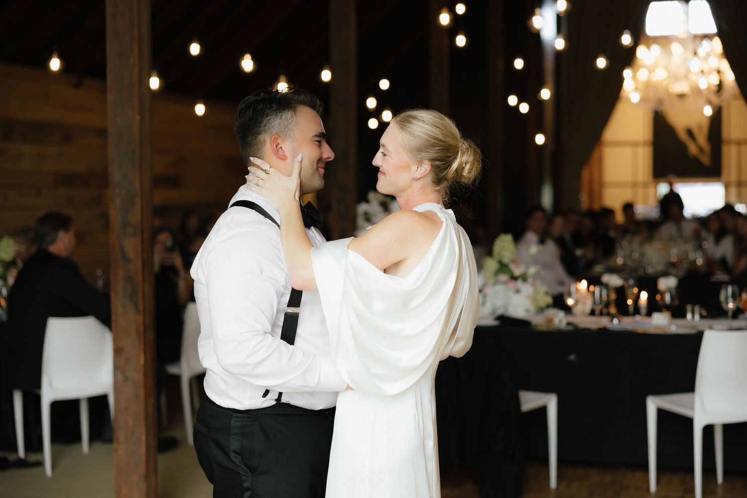Bride and groom sharing first dance in rustic barn venue with warm string lights overhead.
