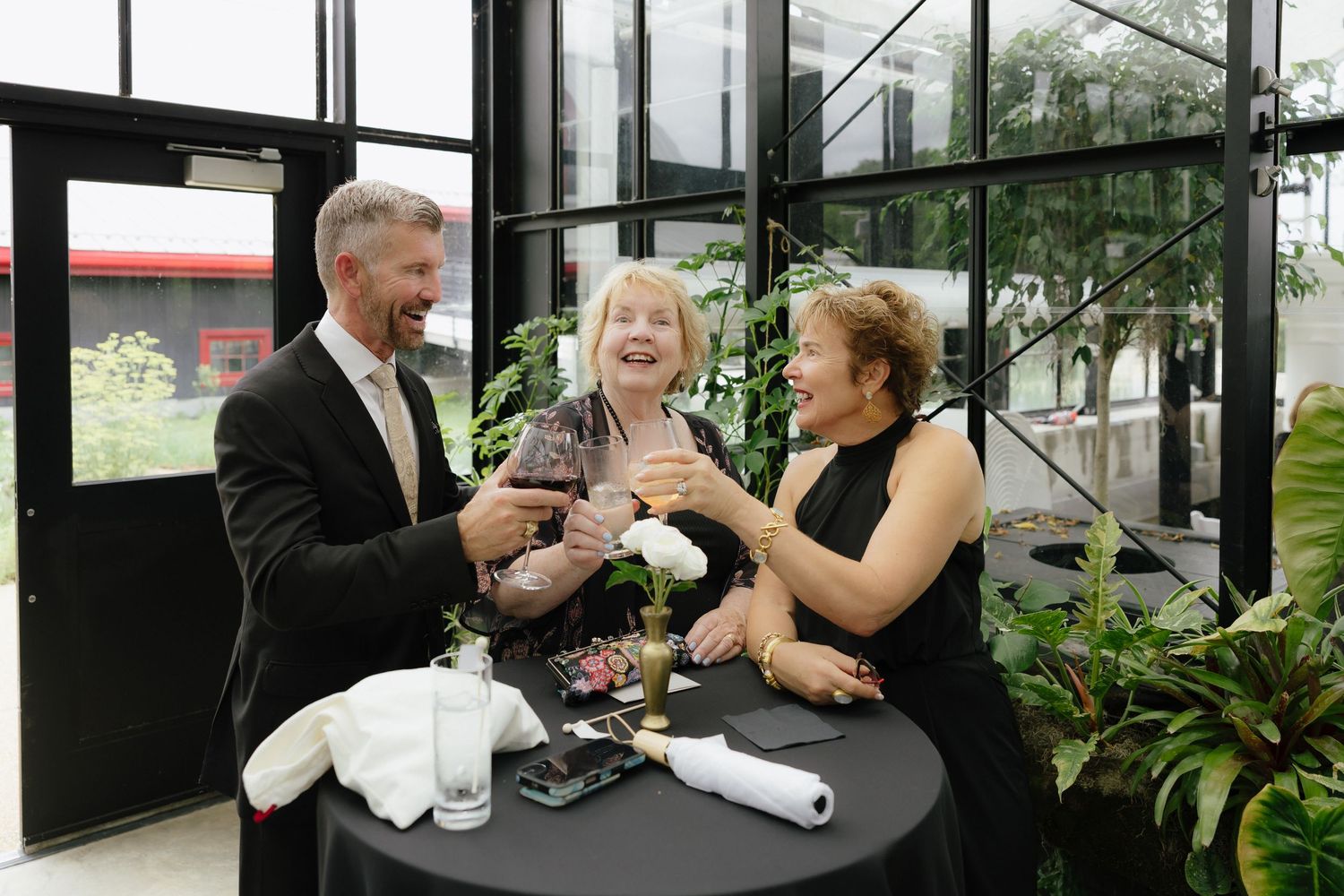 Three wedding guests laugh and raise glasses in a toast at an indoor reception with green plants.