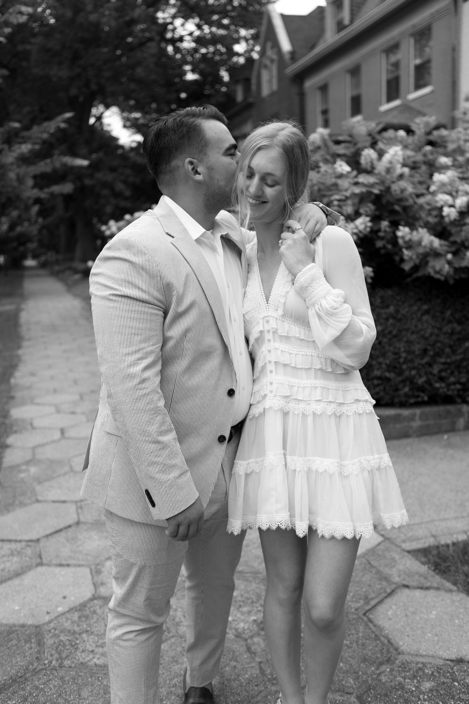 Black and white photo of a couple, man in suit kissing woman in white dress, posing outdoors near blooming flowers.