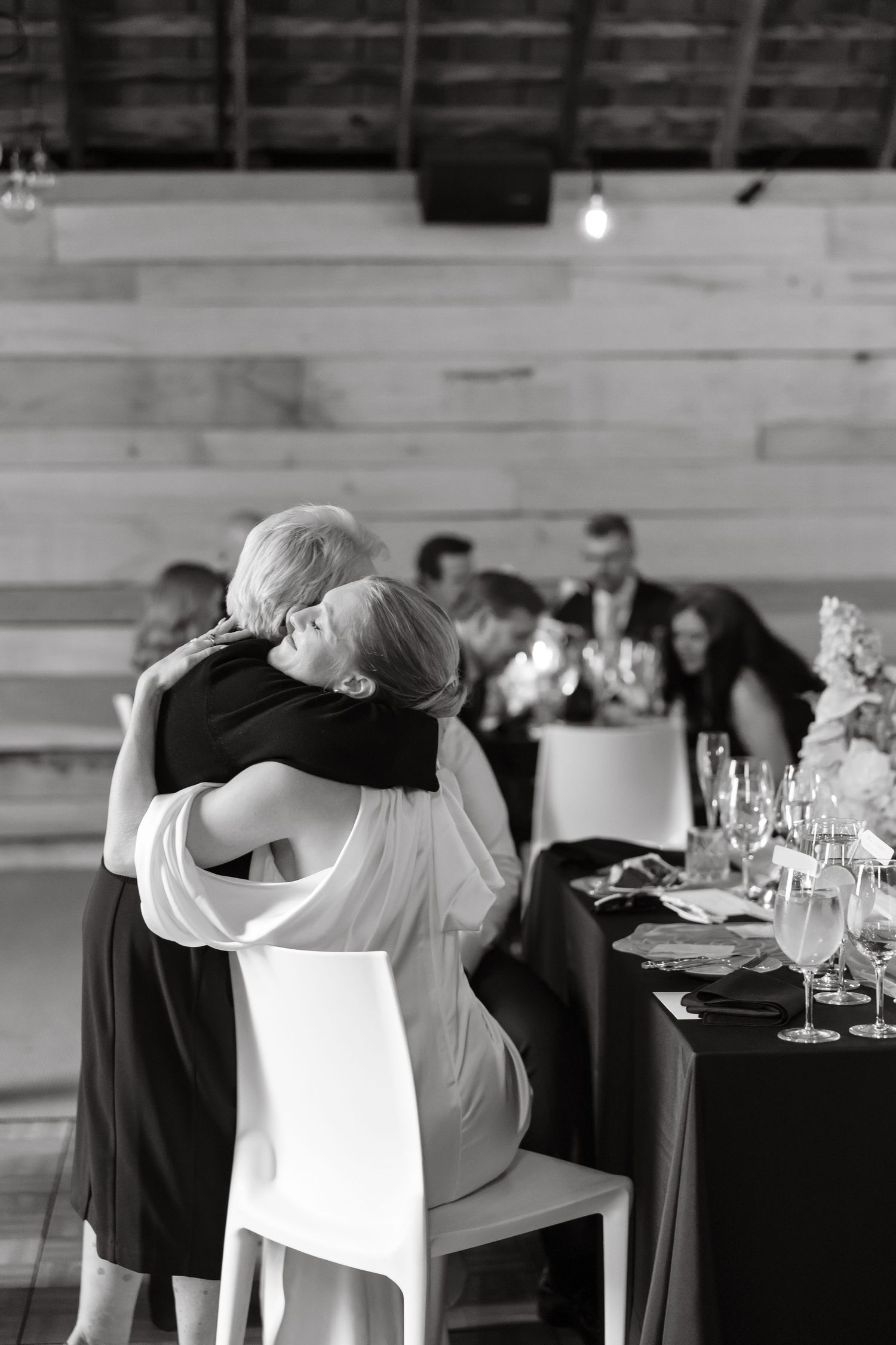 Black and white photo of bride embracing seated elderly woman at wedding reception dinner table.