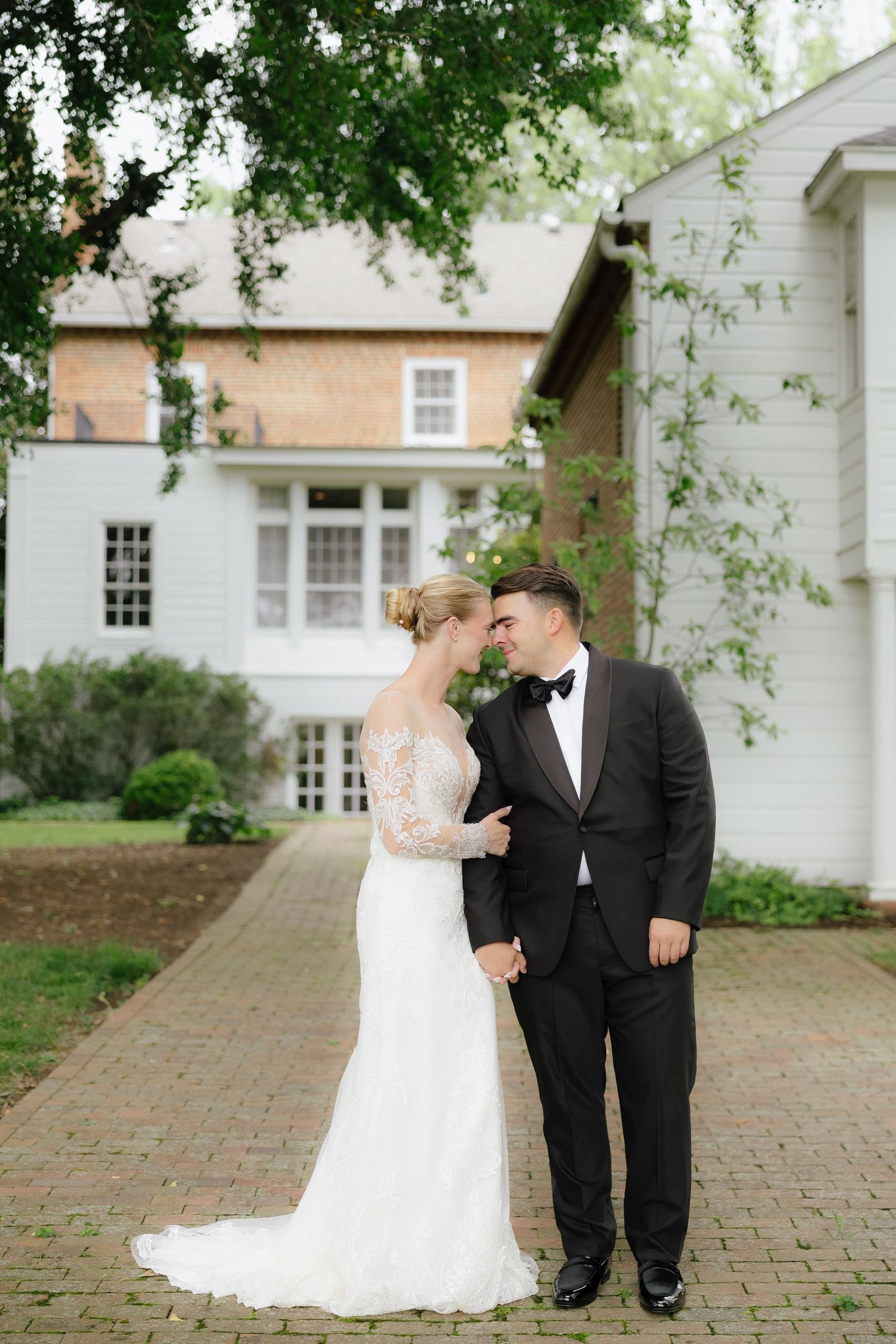 Bride in lace gown and groom in black tuxedo share a romantic moment outside a white colonial wedding venue.