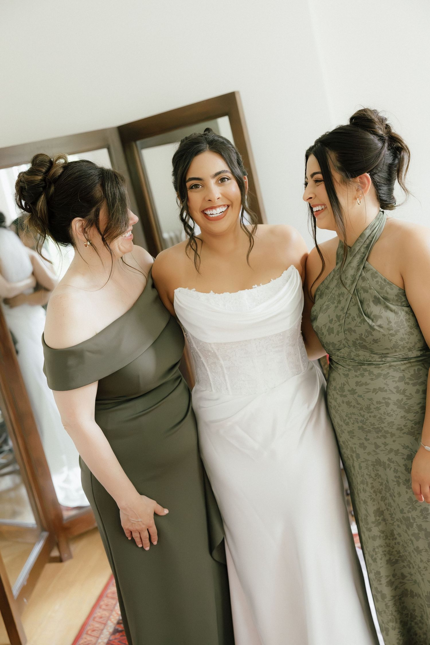 Bride in white strapless gown smiles with two bridesmaids in olive green dresses before wedding ceremony.