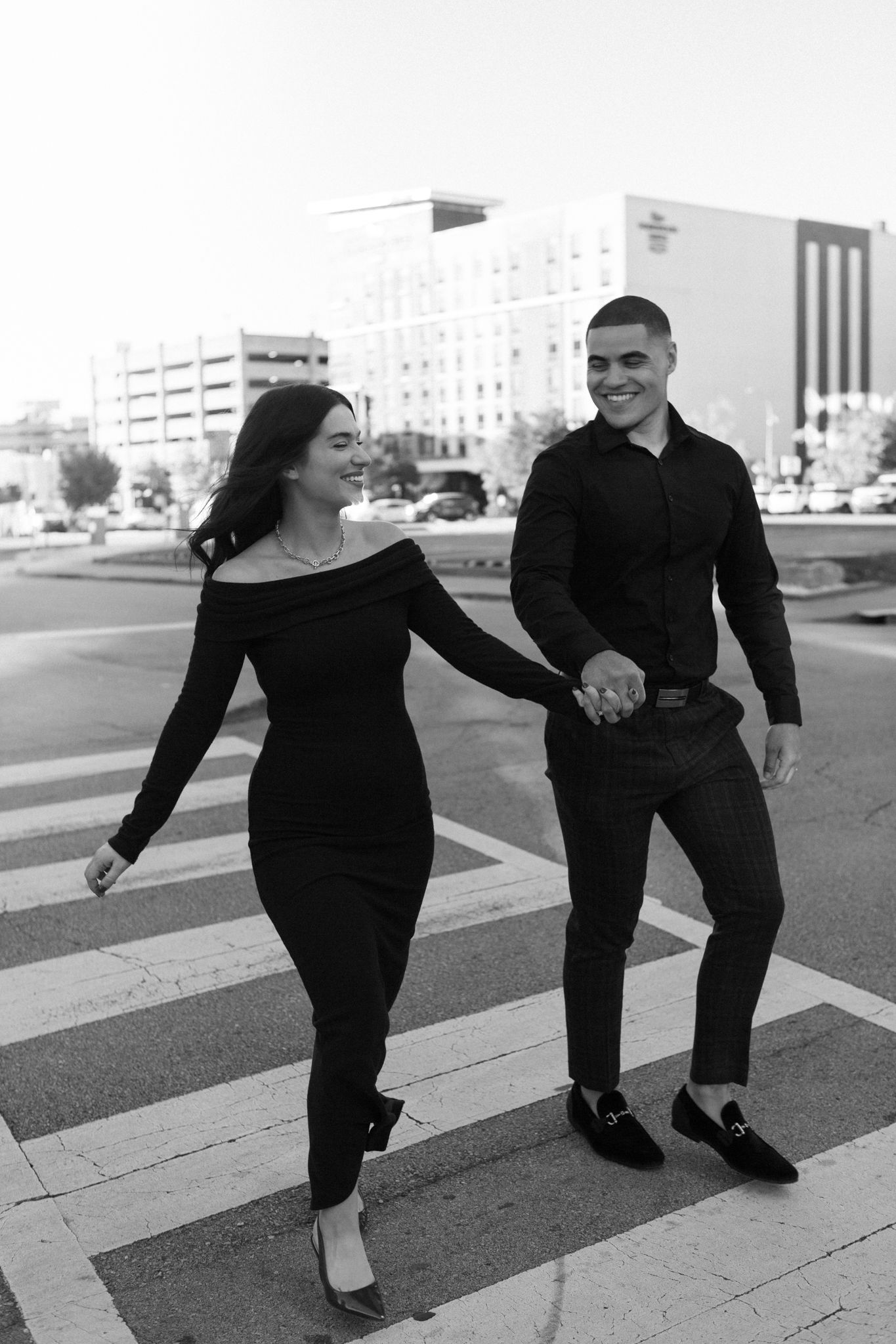 A couple walking hand in hand across a city crosswalk wearing matching black outfits in black and white photography.