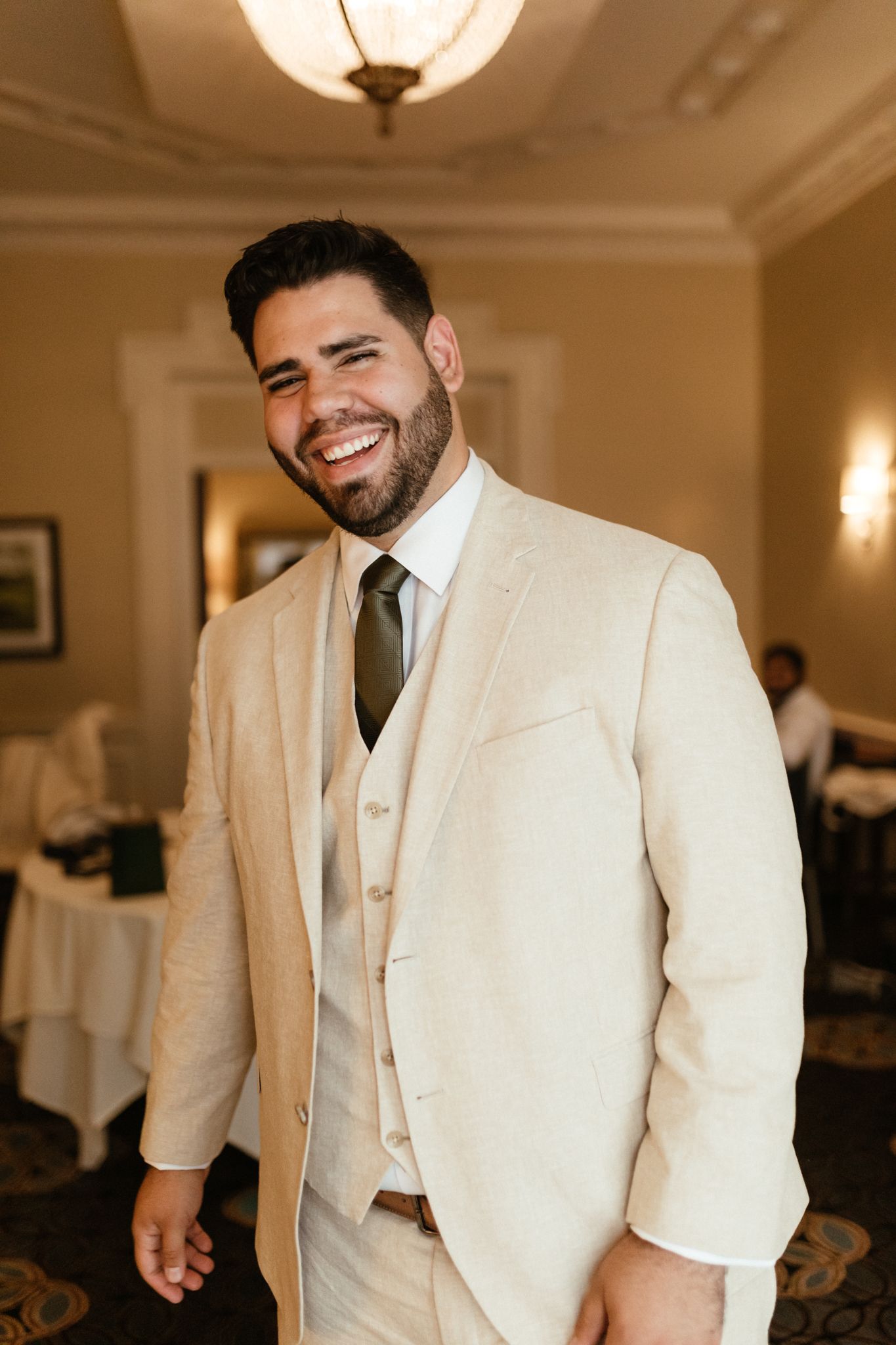 Someone wearing a cream colored three-piece suit and olive tie smiles warmly in an elegant indoor venue.