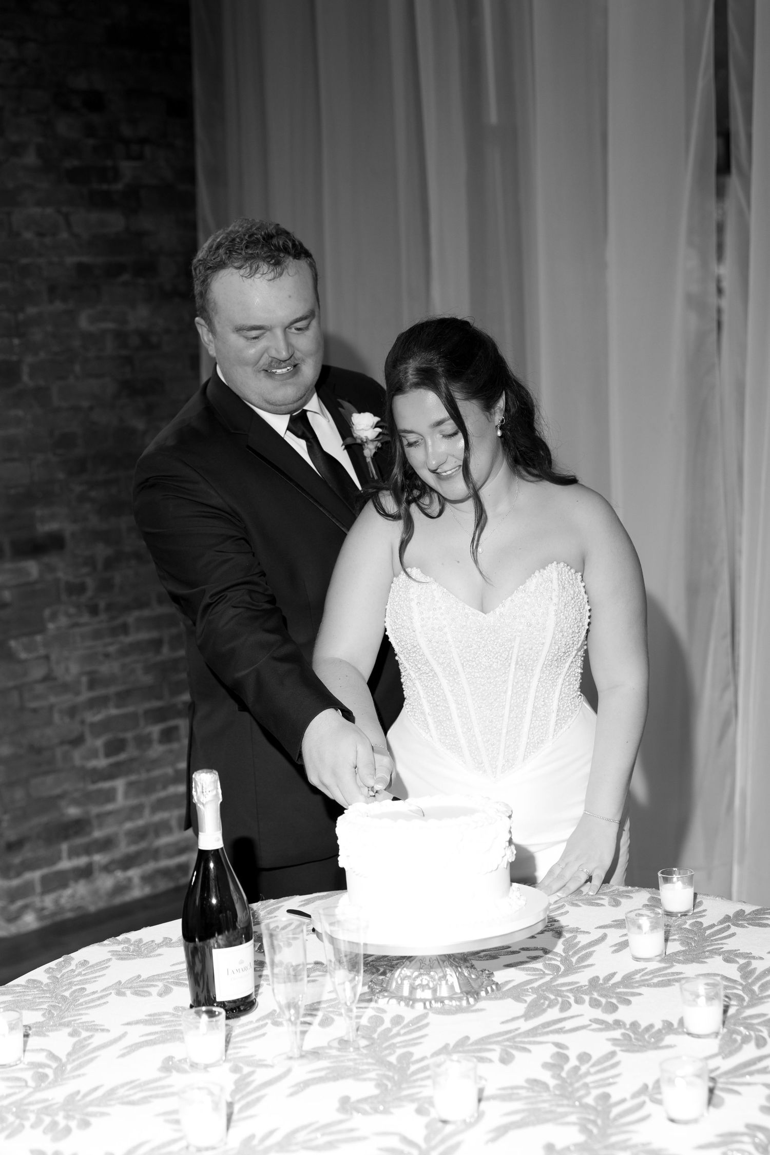 Black and white photo of bride and groom cutting wedding cake together at reception table with champagne bottle nearby.