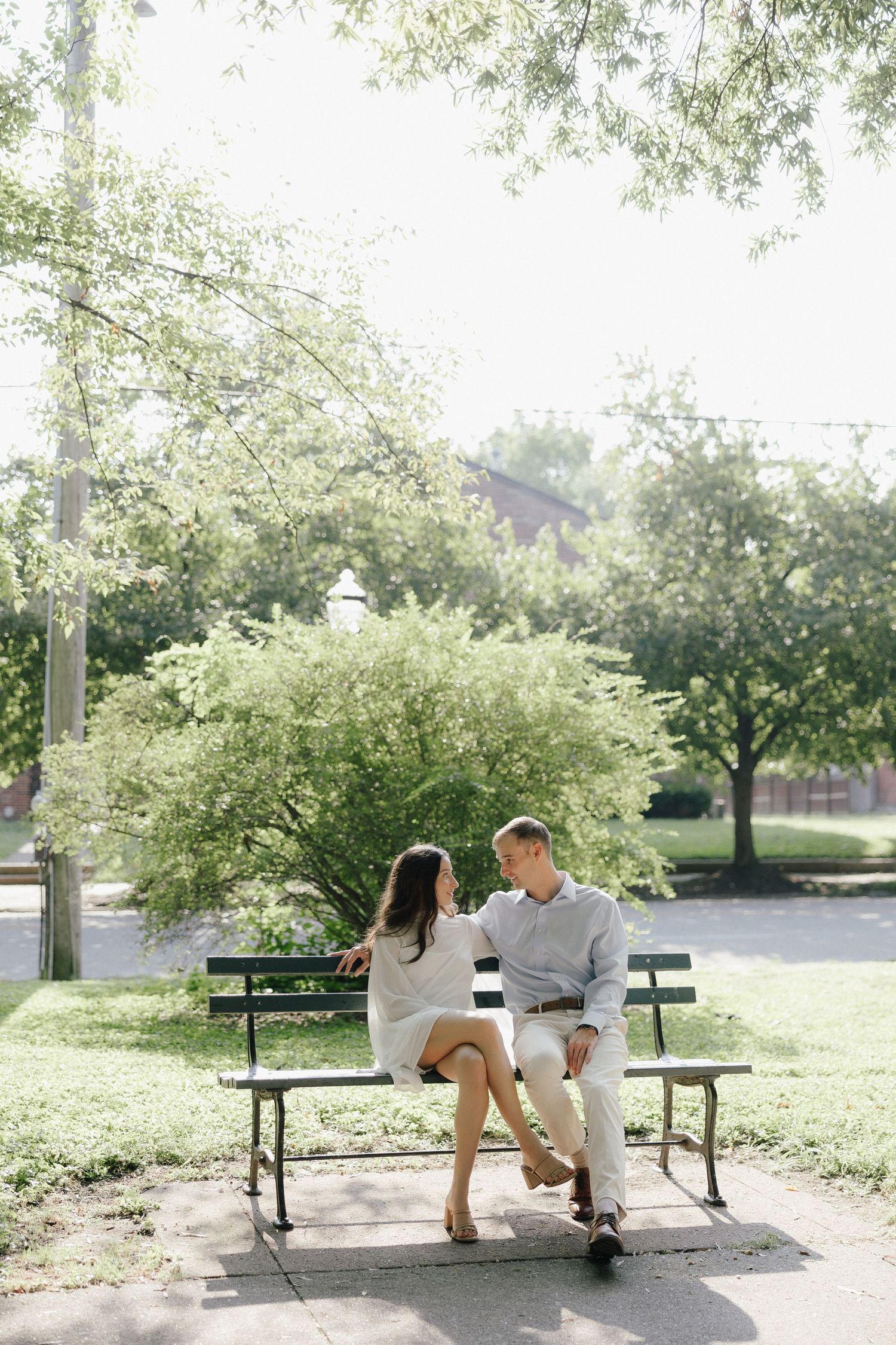 Couple sitting together on a park bench surrounded by lush green trees on a sunny day.