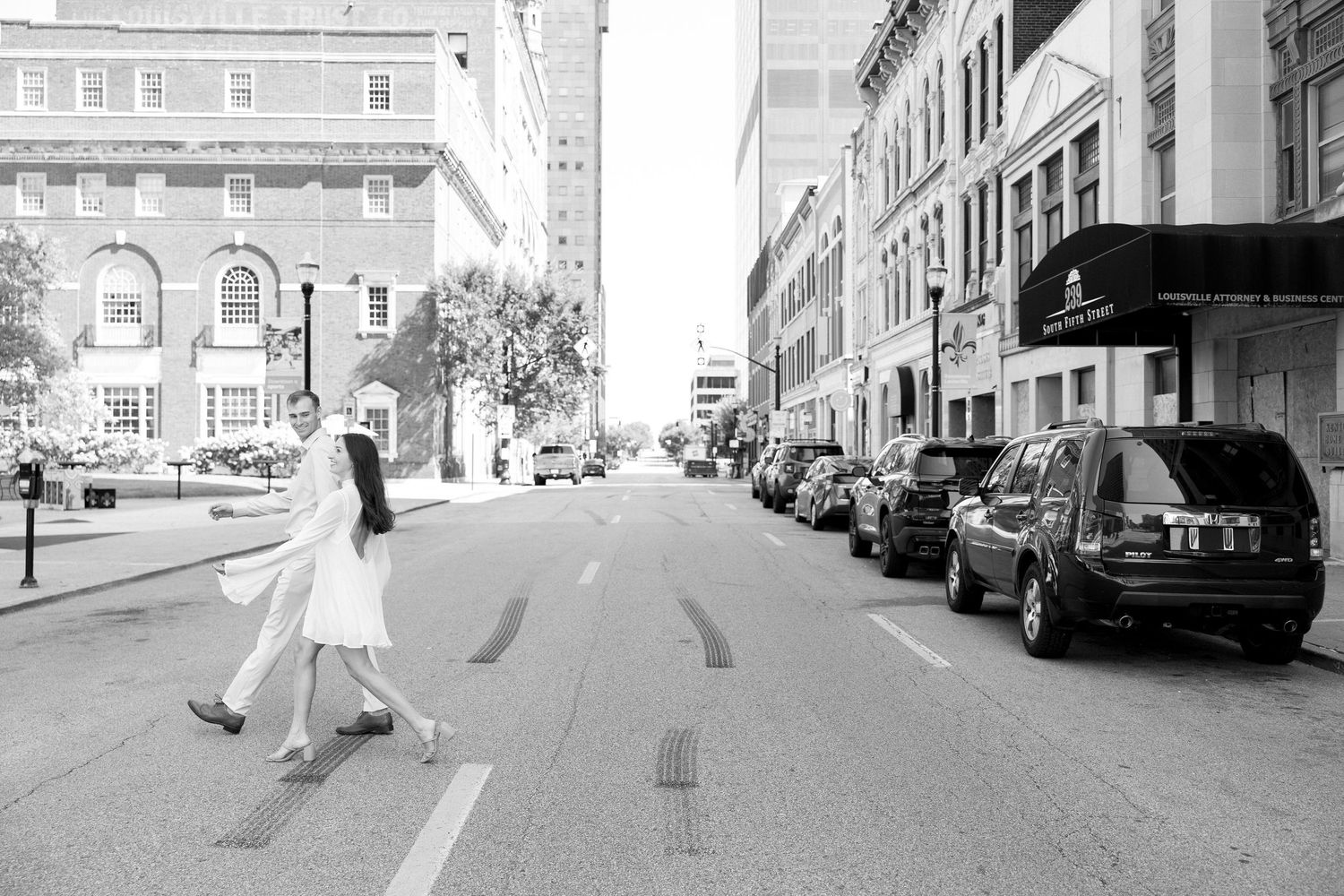 Black and white photo of couple crossing a city street, woman in white dress, urban background.
