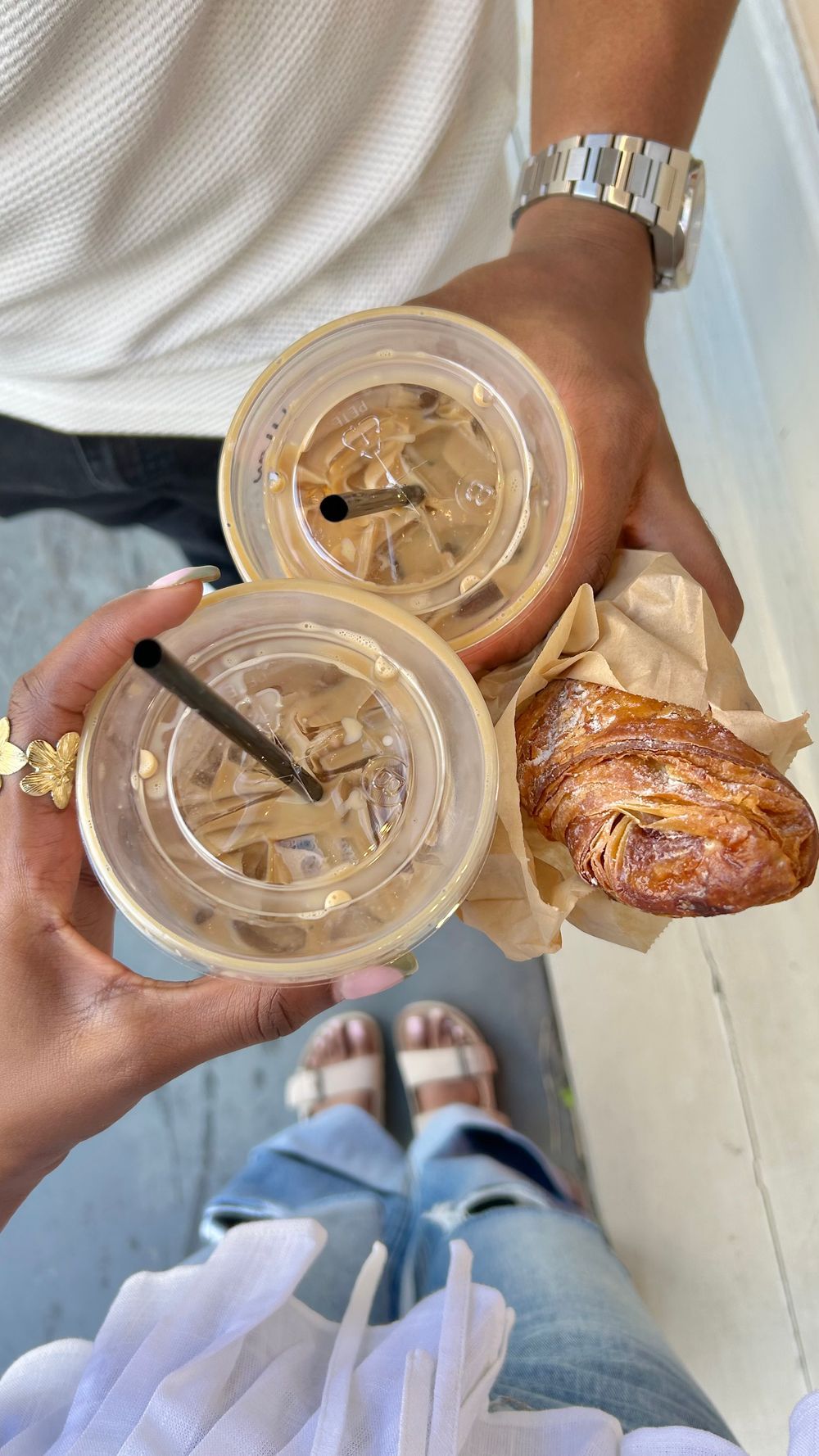 Two iced coffees with plastic lids and a golden croissant wrapped in parchment paper held by two people outdoors.