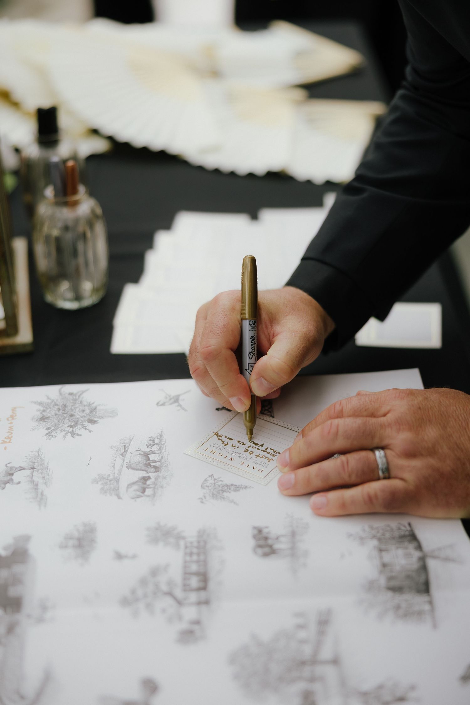 Calligrapher writing elegant script on illustrated wedding stationery at a black table with envelopes.
