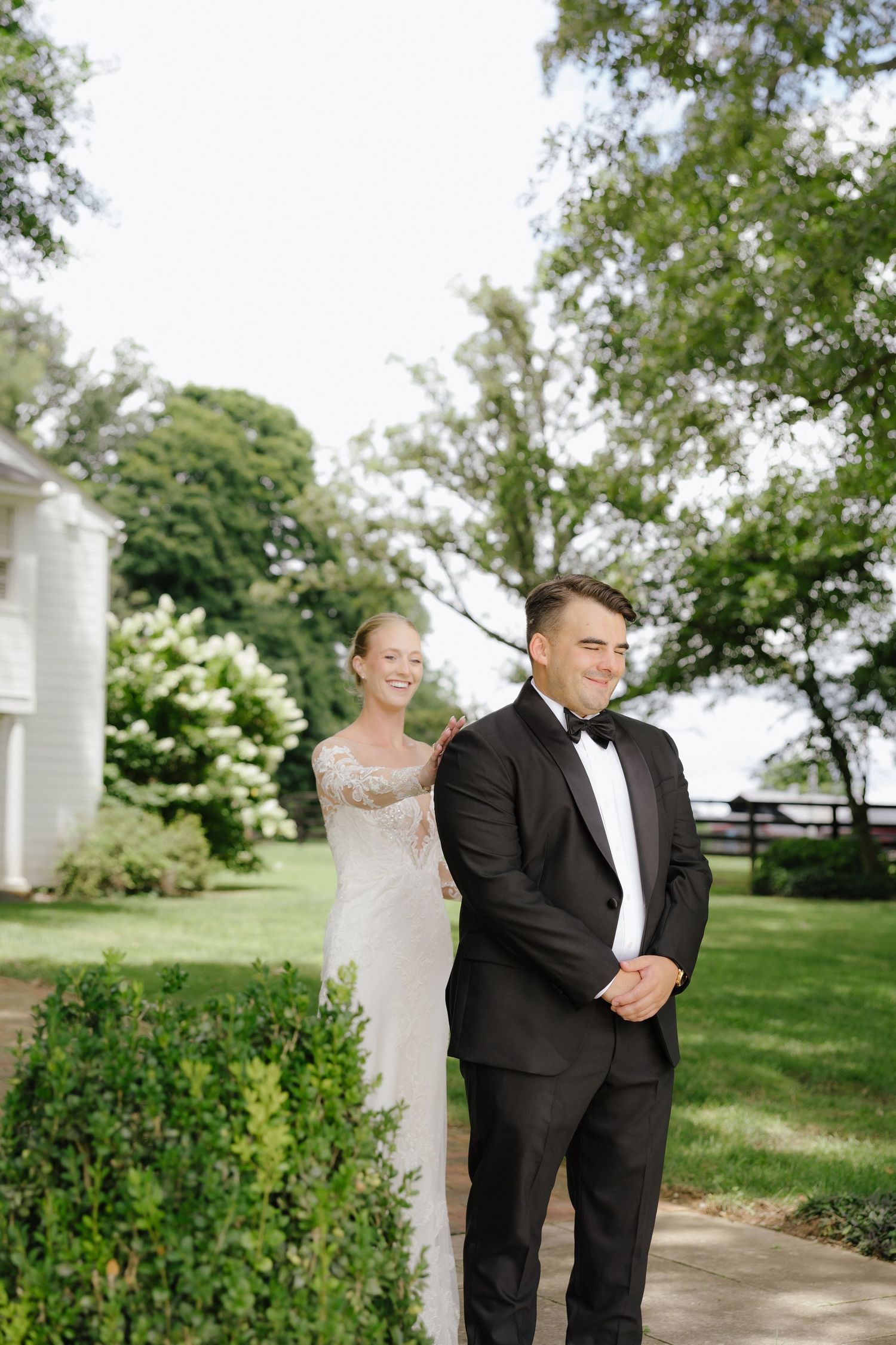 Bride in lace gown surprises groom in tuxedo during outdoor first look at garden wedding venue.