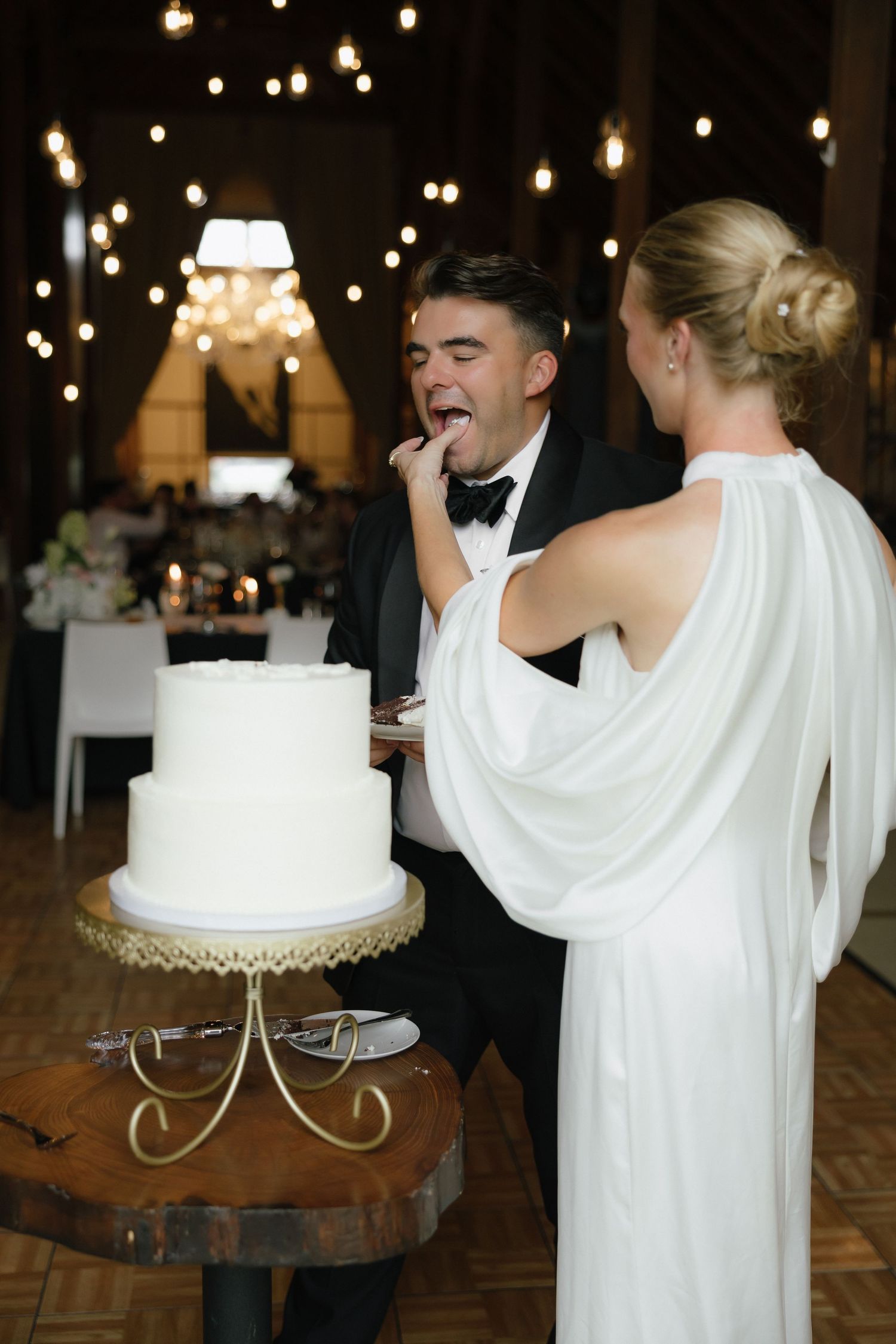 Bride in white gown feeds groom wedding cake at rustic reception venue with warm string lights.