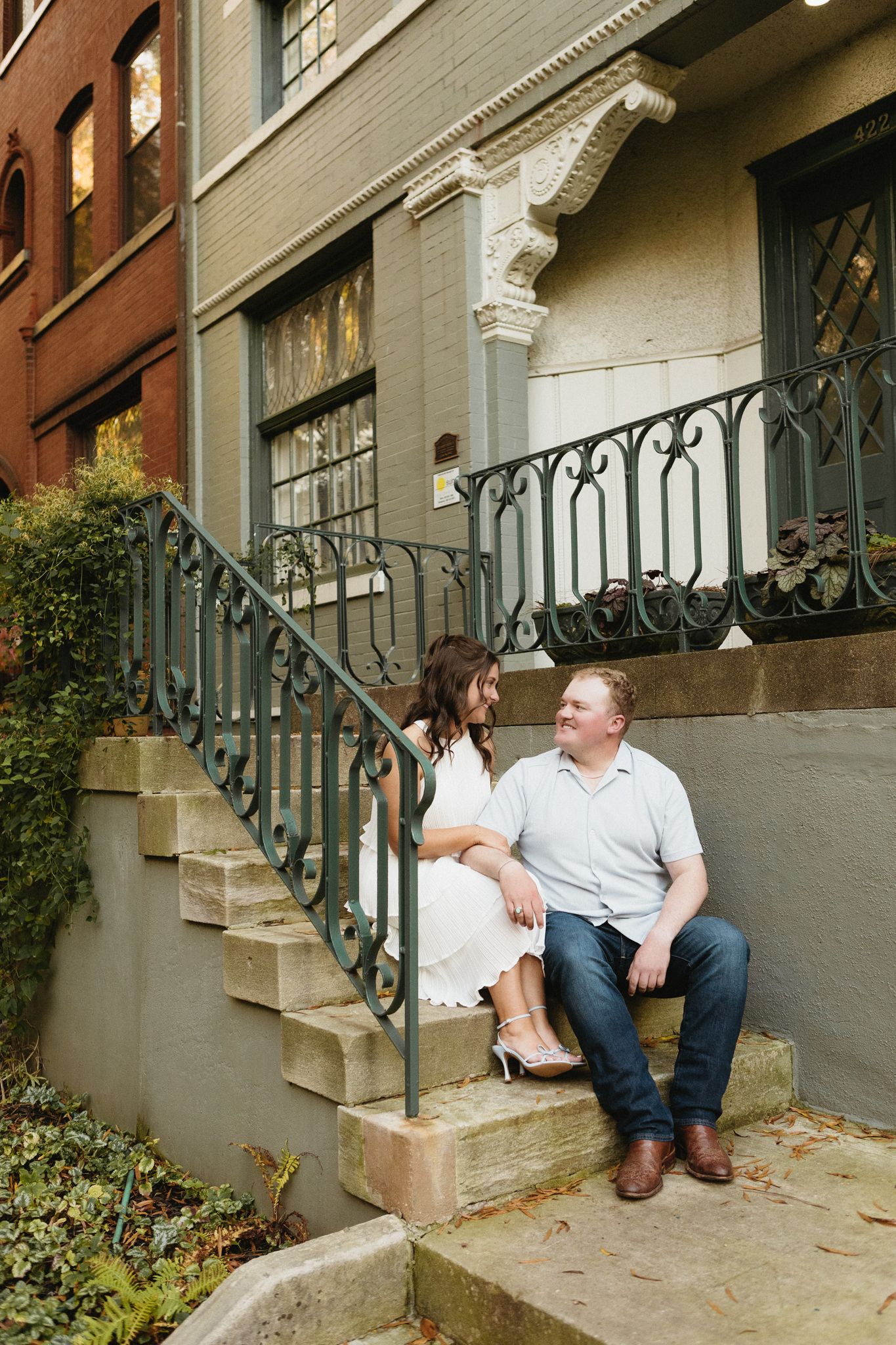 A couple sits together on stone steps with ornate iron railings outside a historic brownstone building.